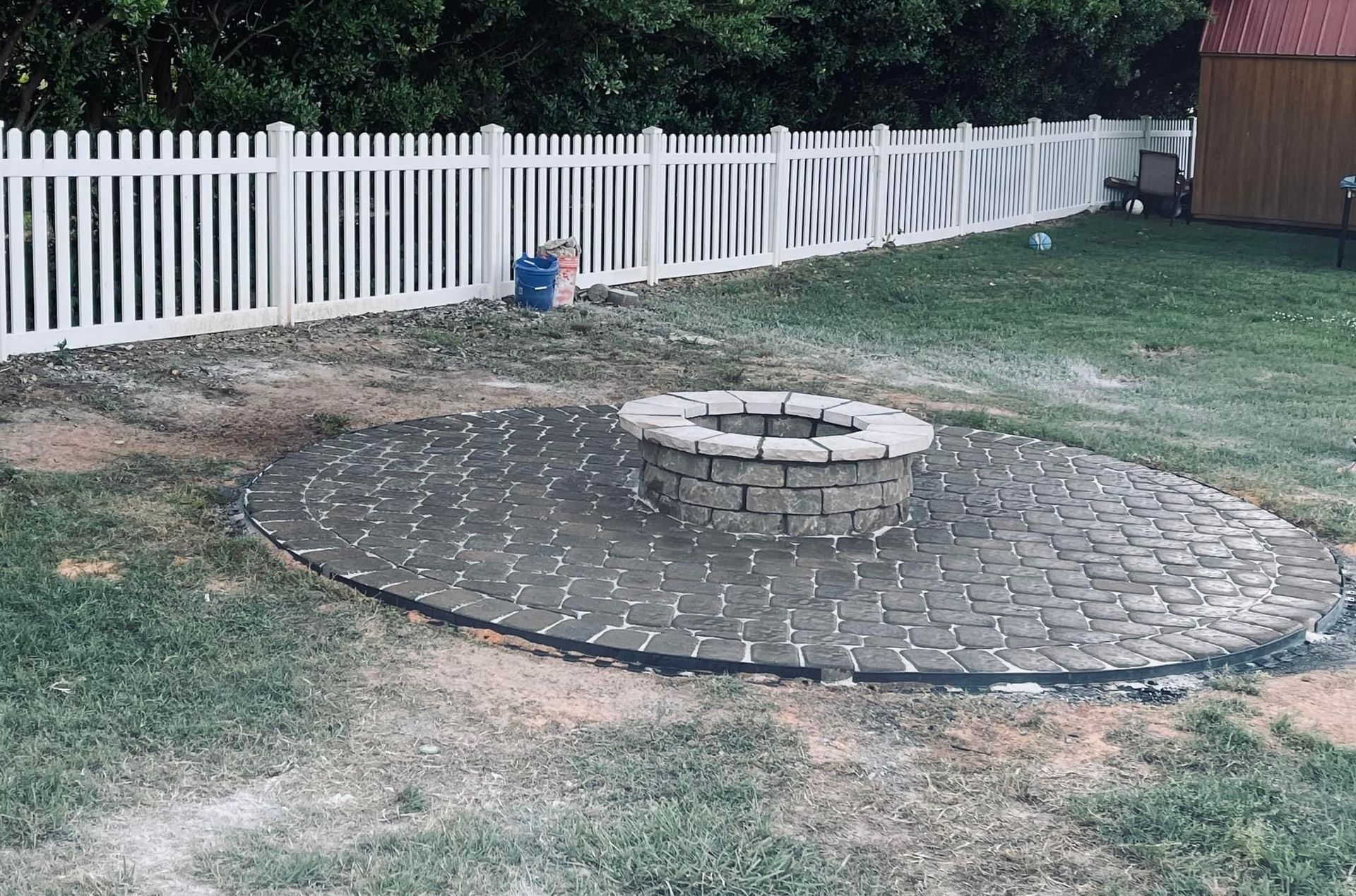 A fire pit made of stone, on a circular stone patio, in a grassy backyard, with a white picket fence.
