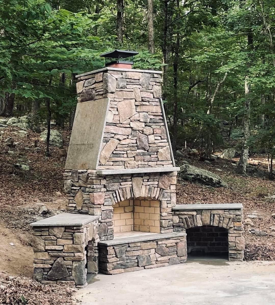 Stone outdoor fireplace with a brick interior, set in a wooded area.