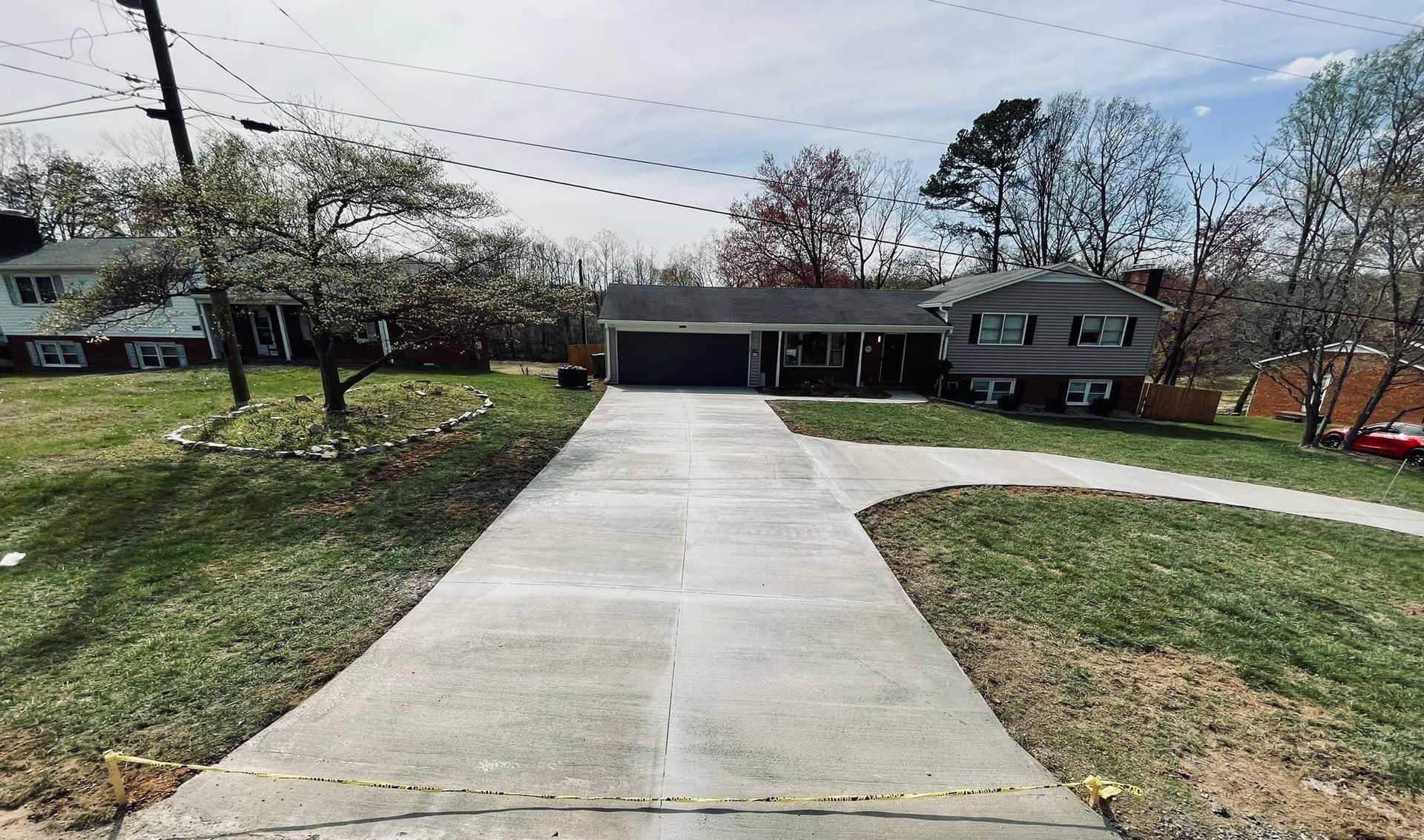Concrete driveway leading to a one-story house with a dark roof, surrounded by green grass and trees.