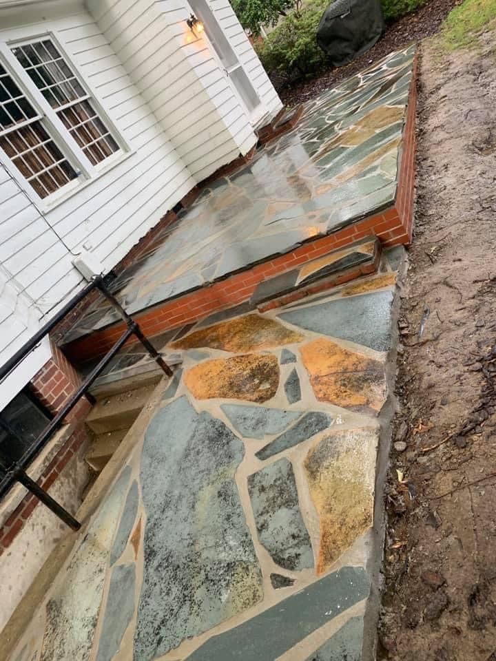 Stone walkway with steps leading to a white house, featuring a brick wall and black handrail.