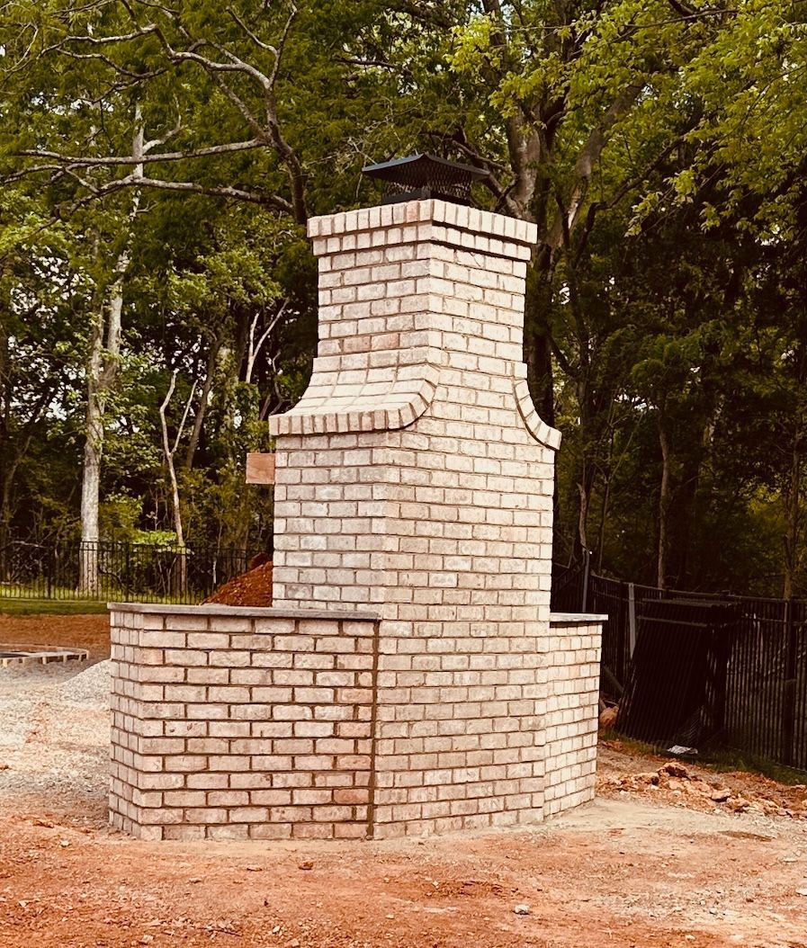 Brick outdoor fireplace with curved sides and a small chimney, set against trees.