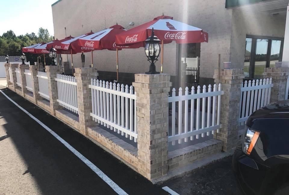 Outdoor restaurant seating area with white picket fence, red Coca-Cola umbrellas, and stone pillars.