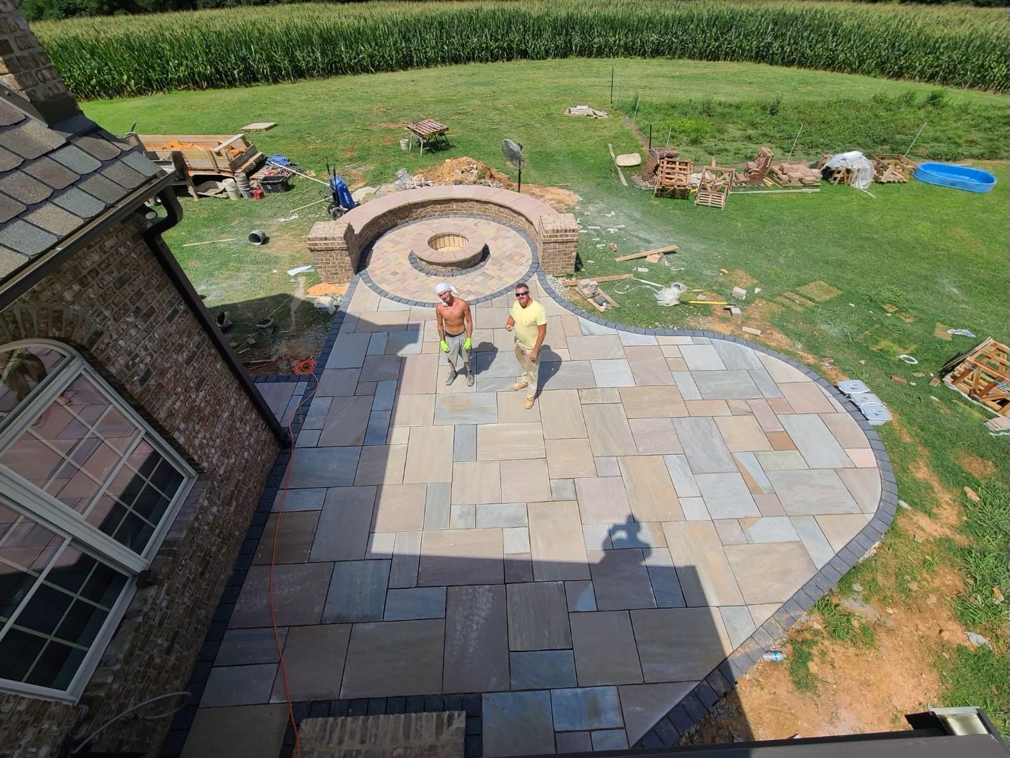 Two men standing on a newly constructed stone patio with a fire pit in the background.