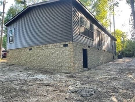A house with gray siding on a stone block foundation; set in a dirt yard surrounded by trees.