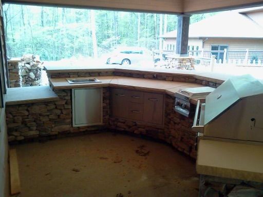 Outdoor kitchen with stone facade, concrete countertop, grill, and a view of trees.