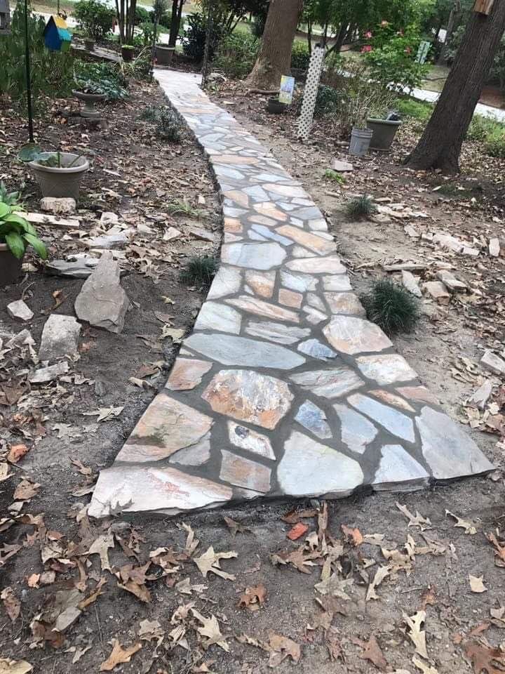 Stone pathway winding through a garden with trees, leaves, and potted plants.
