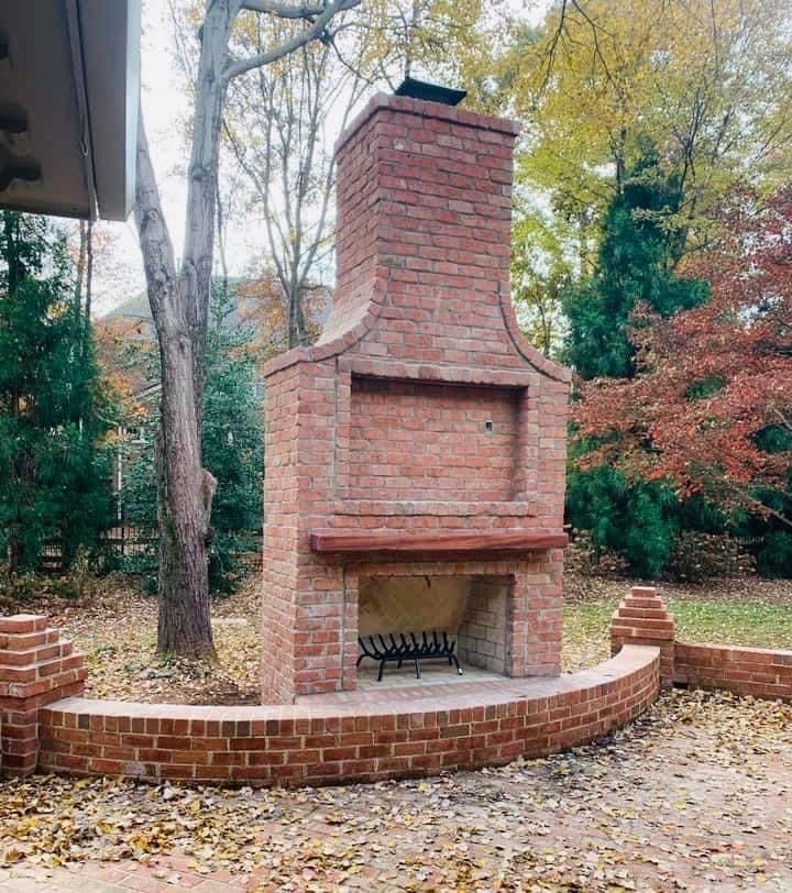 Brick outdoor fireplace with arched front and a curved brick seating wall in a backyard with autumn leaves.