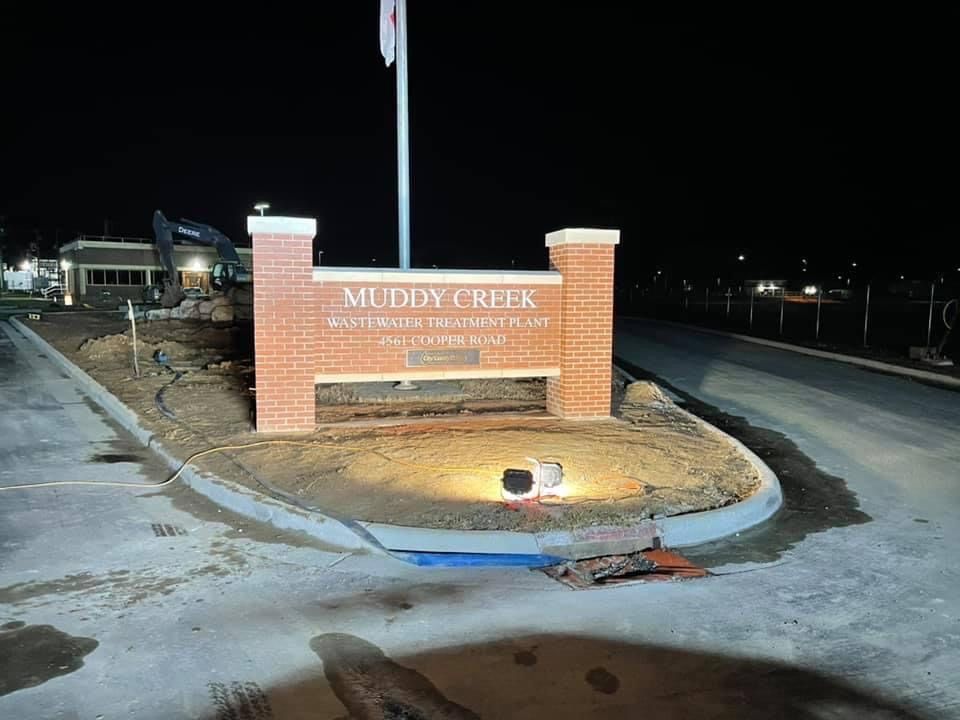 Muddy Creek entrance sign at night with brick pillars and flag, lit by a spotlight.
