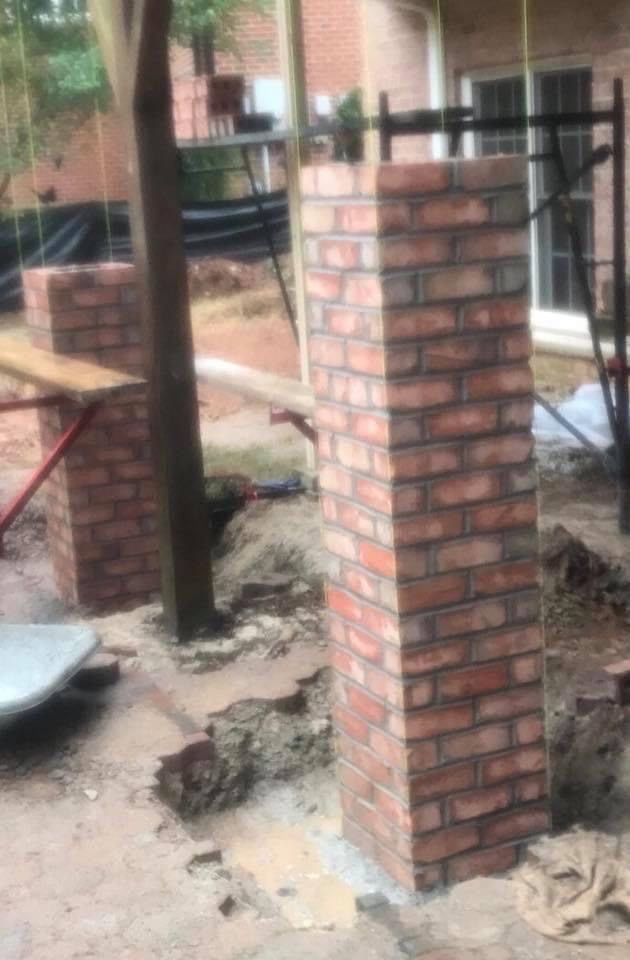 Brick columns under construction outside. Red bricks being laid; dirt and tools present.