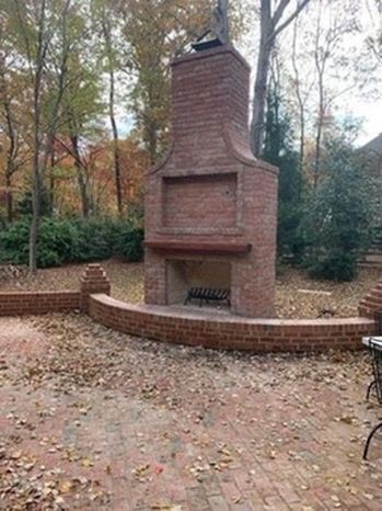 Outdoor brick fireplace with brick patio, surrounded by trees and bushes, autumn leaves on ground.