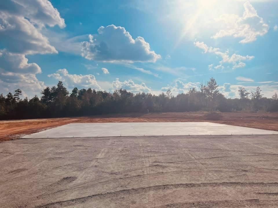 Sunny day view of a concrete pad in a dirt field, trees in the background, blue sky with clouds.
