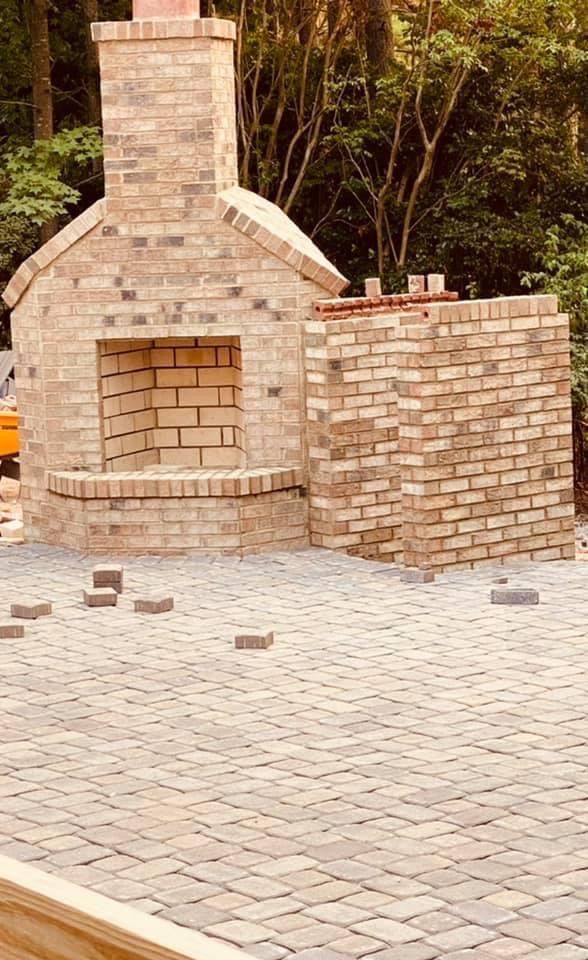 Brick outdoor fireplace under construction on a paved patio in a wooded setting.
