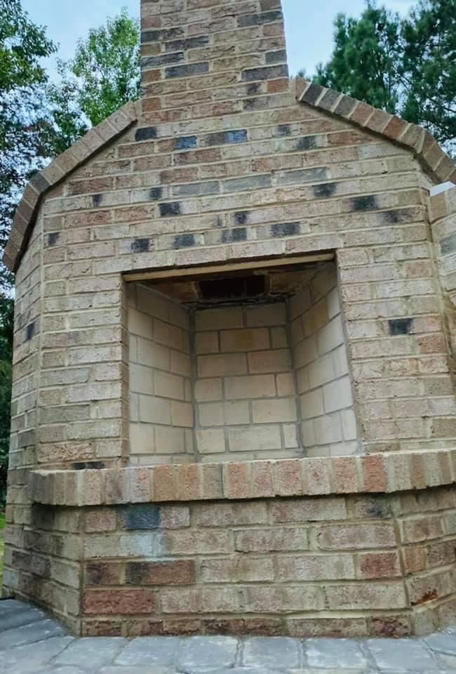 Brick outdoor fireplace, beige and brown brick, with an open firebox, in a wooded setting.