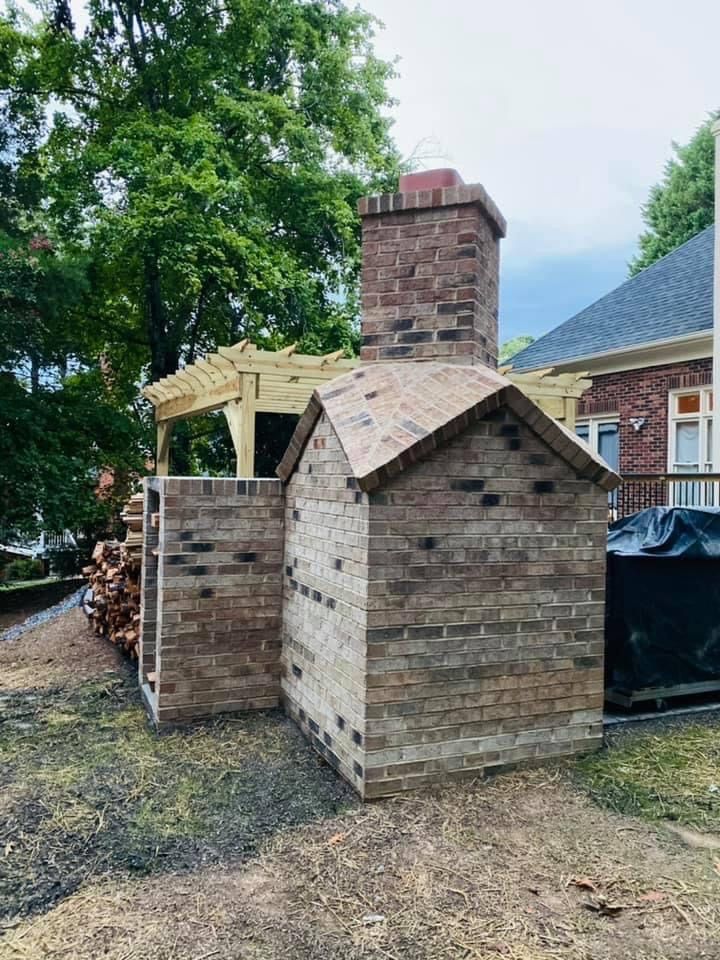 Brick outdoor fireplace with a chimney, next to a wooden pergola and a house.
