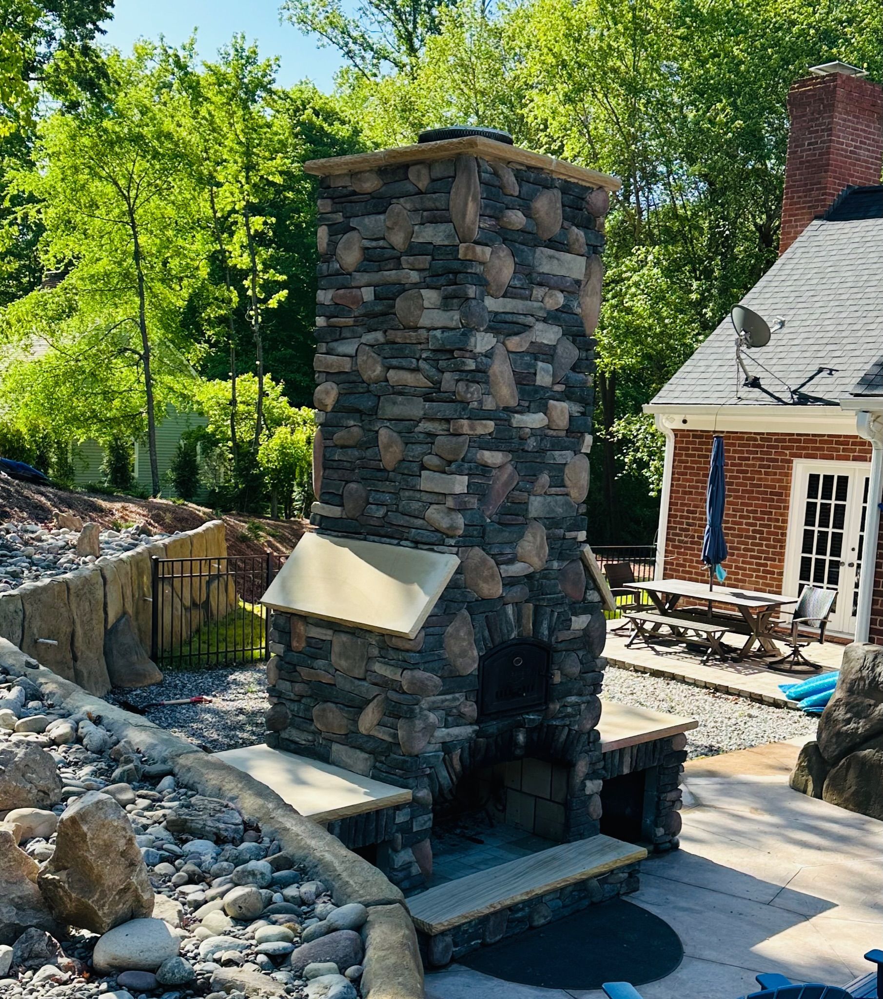 Stone outdoor fireplace with a chimney on a patio with landscaping and a house.