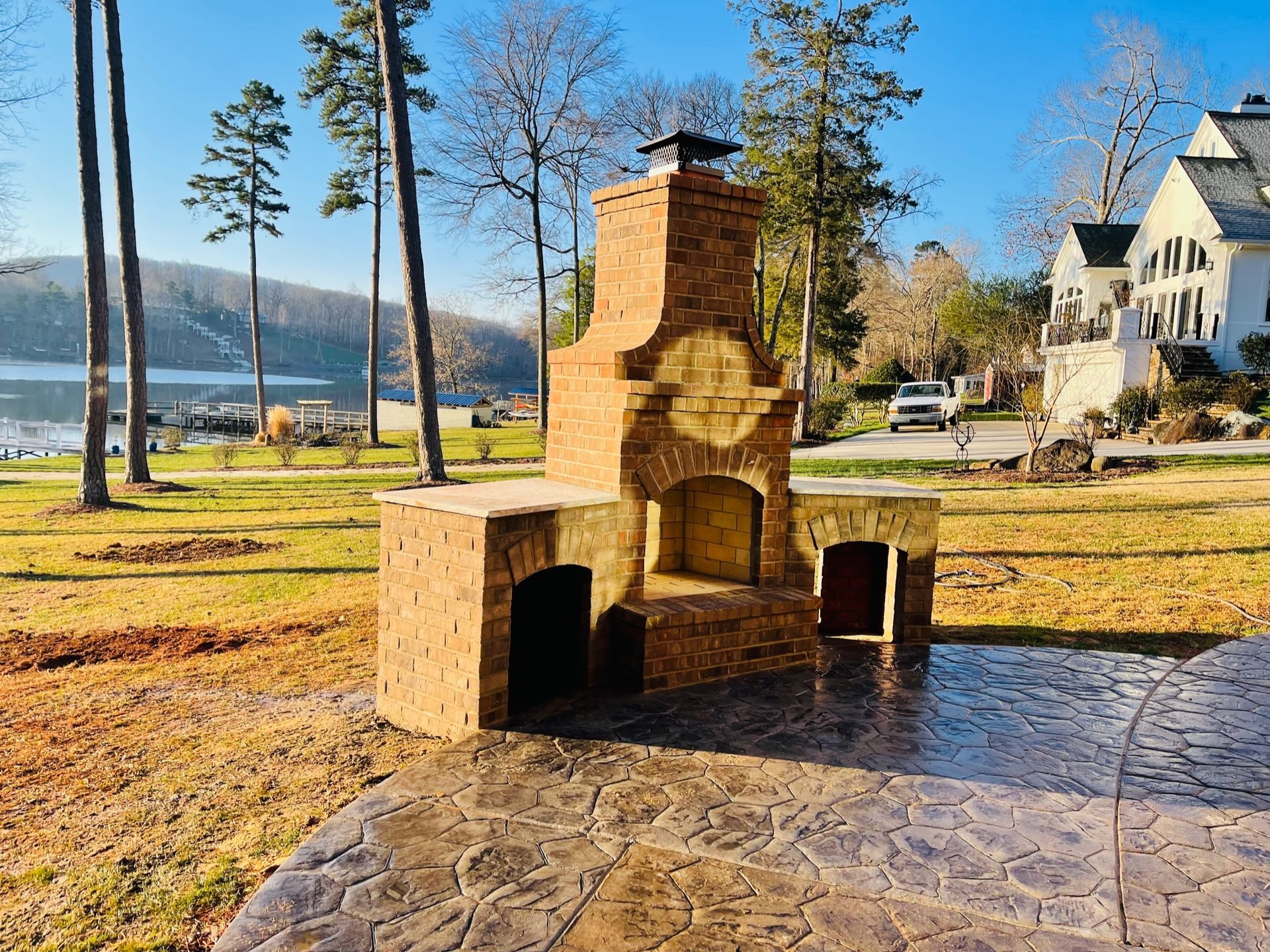 Brick outdoor fireplace and cooking area on a stamped concrete patio, near a lake and trees.