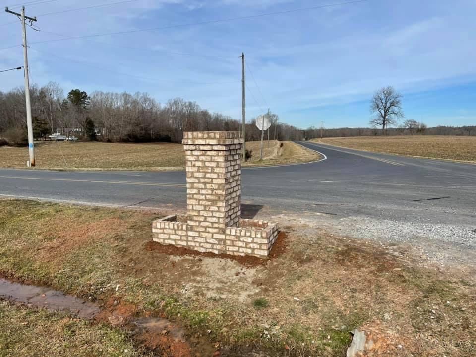 Brick pillar and sign at a road intersection in a rural setting; blue sky, bare trees.