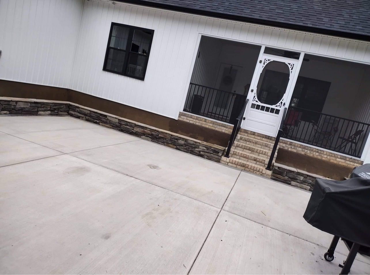 Concrete patio with stone ledge, white house with screened porch, and grill.