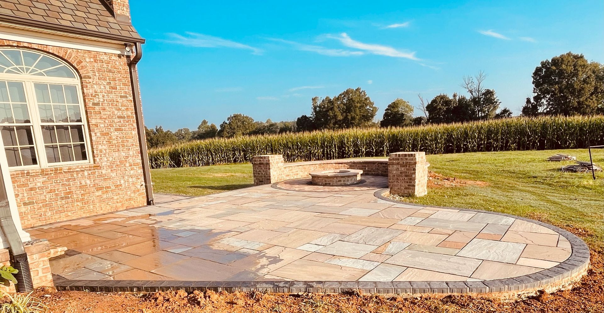 Stone patio with fire pit next to a brick house and a field, under a blue sky.