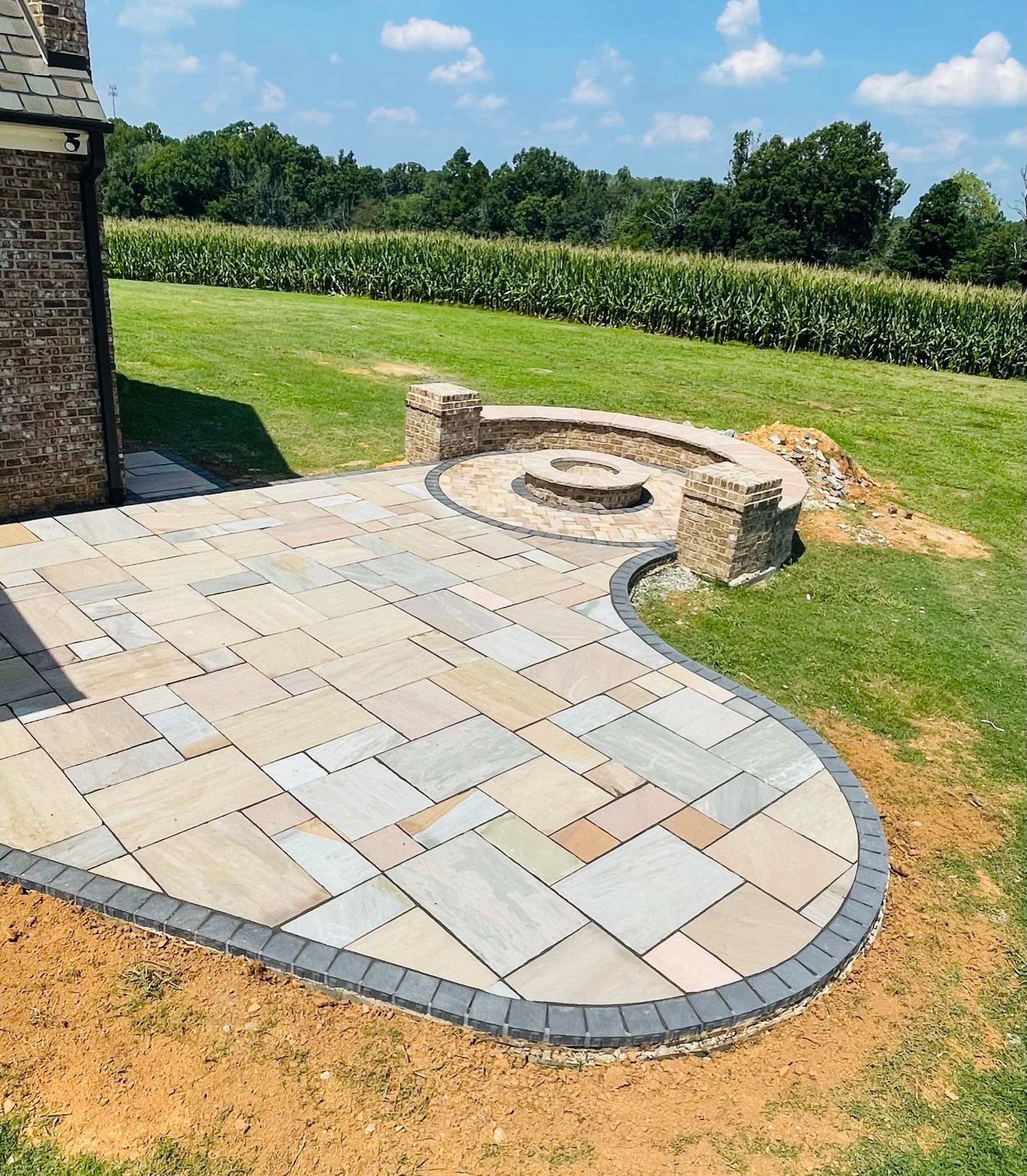 Stone patio with fire pit, brick edging, and a field backdrop on a sunny day.
