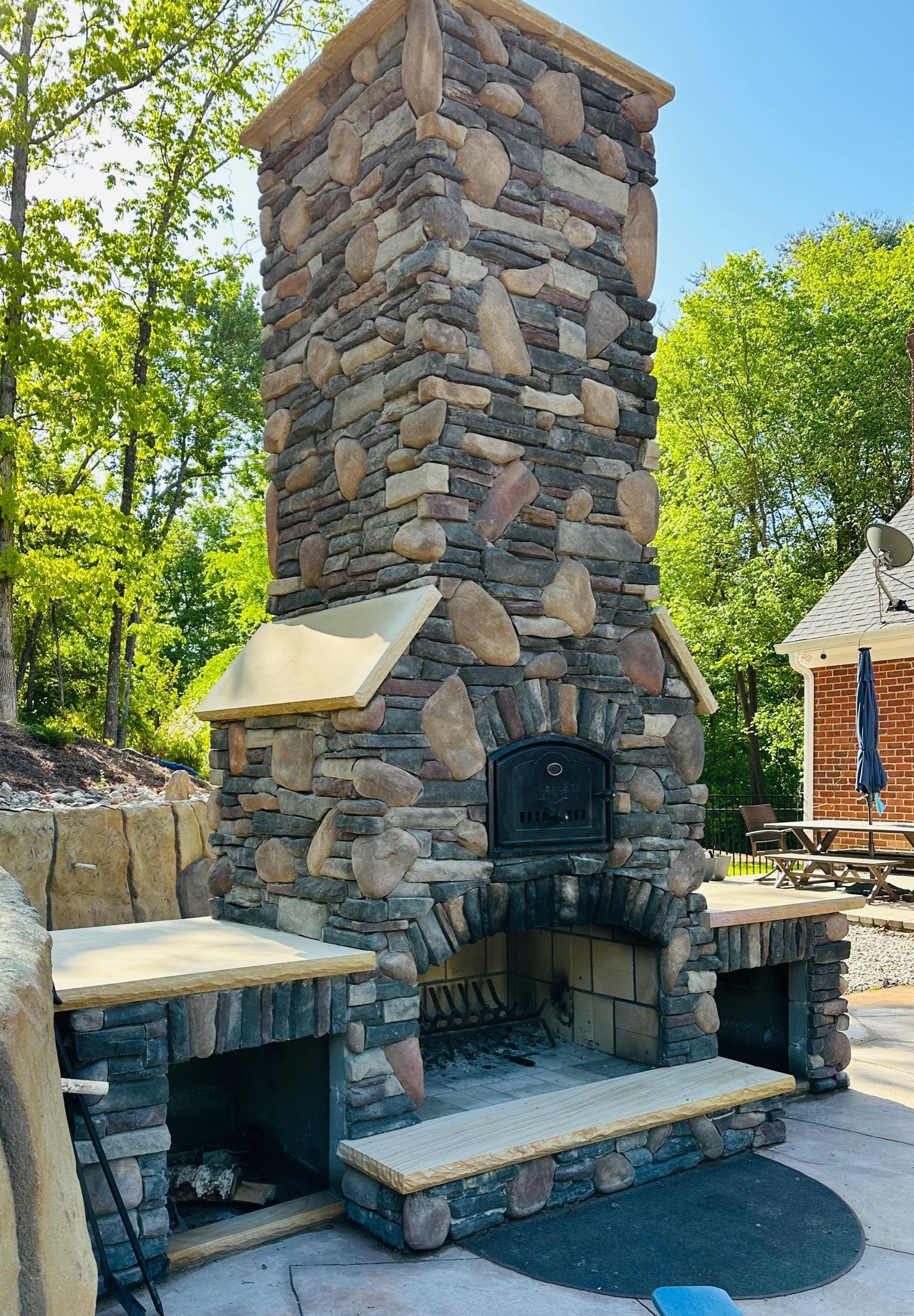 Stone outdoor pizza oven with chimney, counters, and surrounding landscaping.