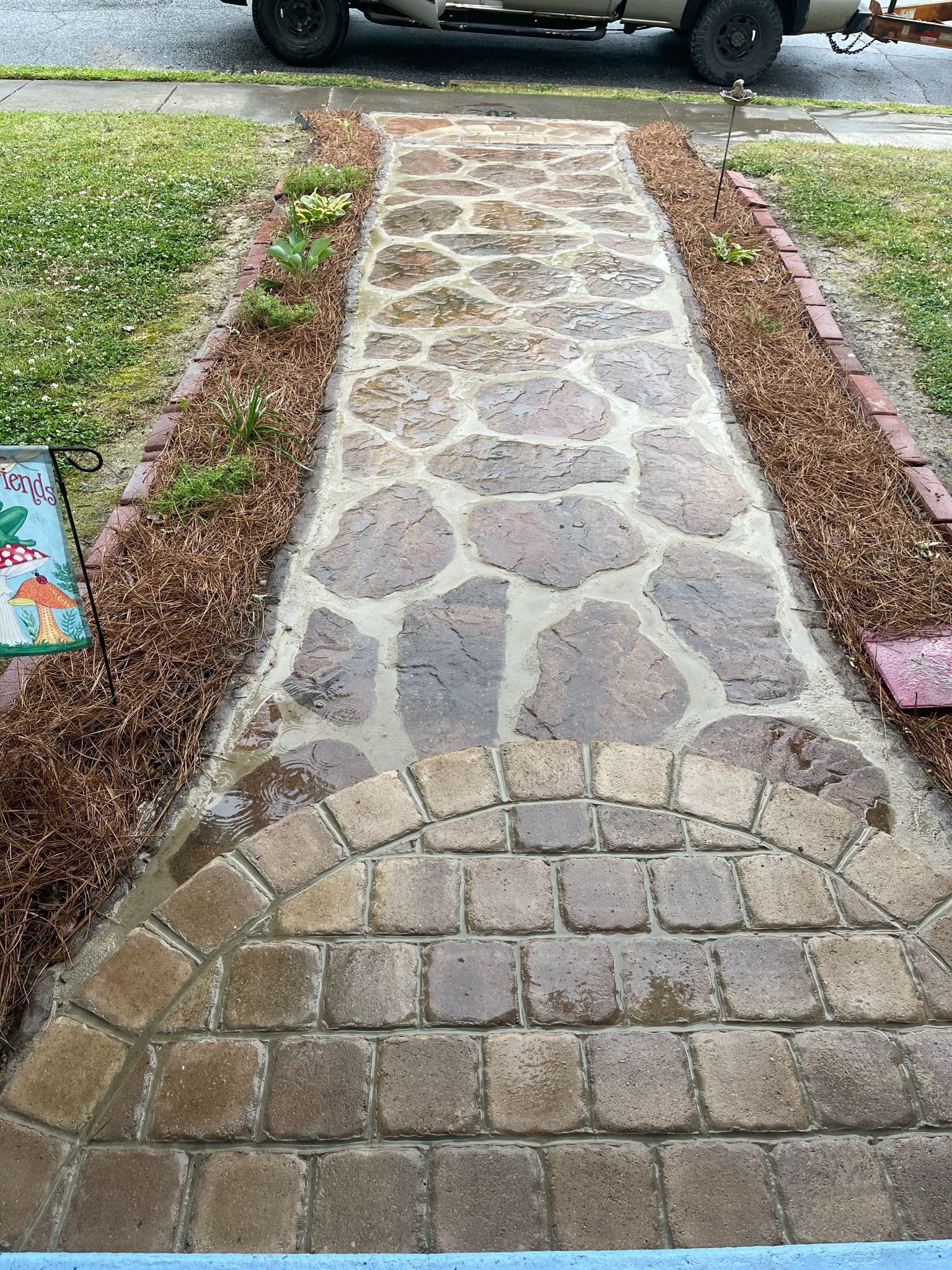 Stone pathway leading to a house, flanked by mulch and grass. The pathway appears wet.