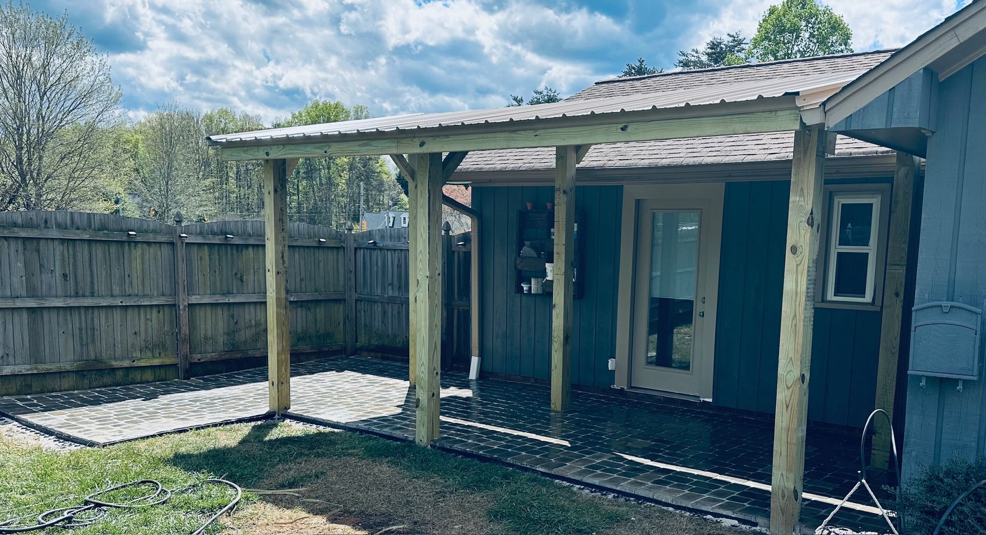 Wooden patio with metal roof attached to a blue house, next to a wooden fence.
