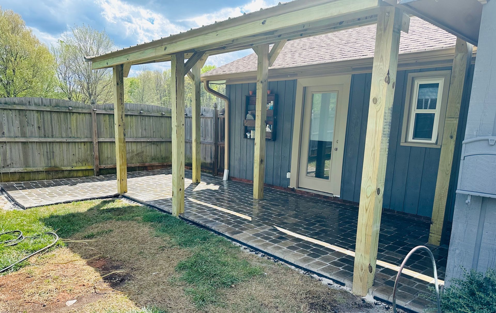 A gray-painted shed with a wood-framed overhang, patio, and fence. Green grass and gravel surround it.