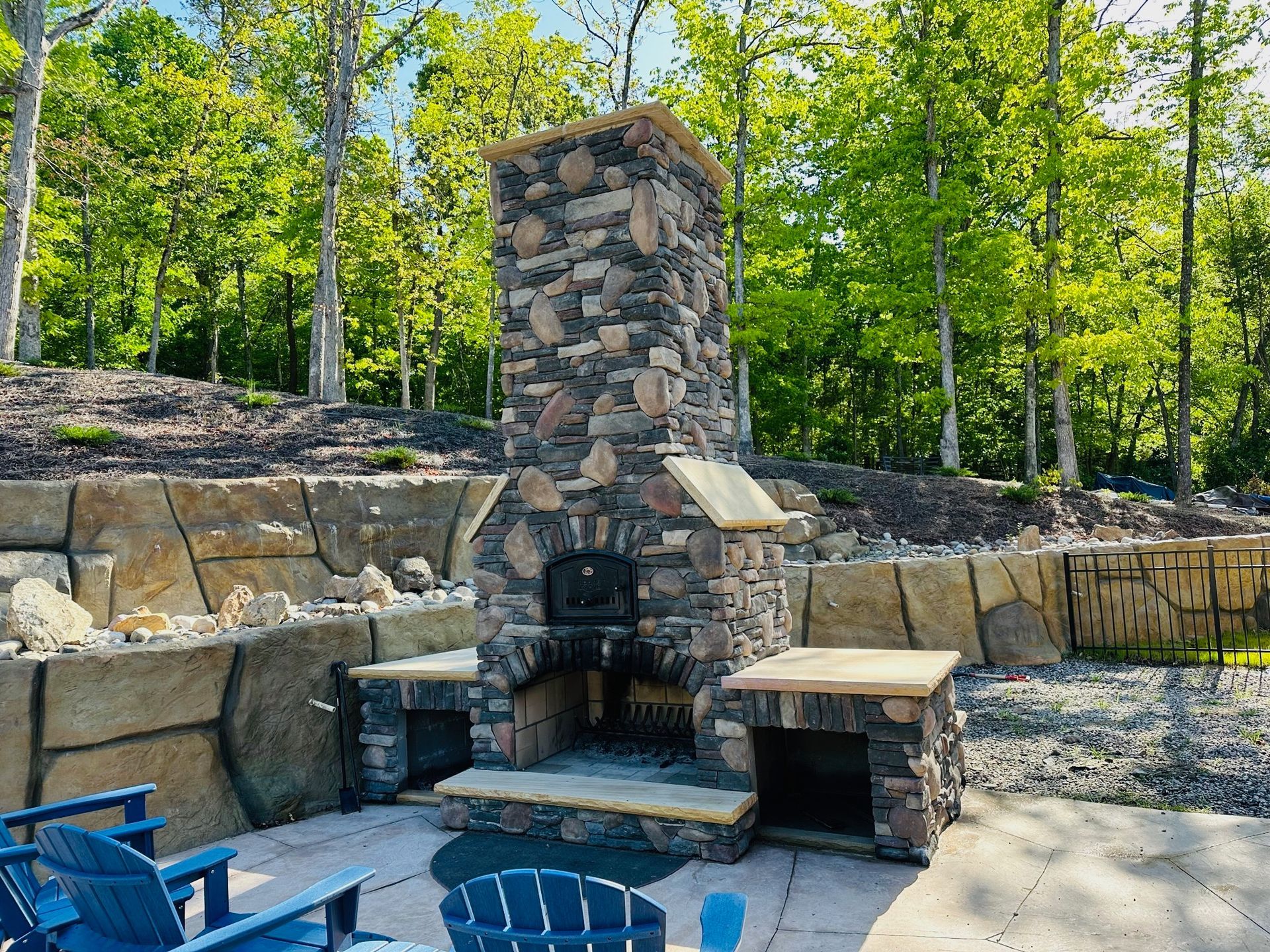 Stone outdoor fireplace with pizza oven, surrounded by patio and trees.