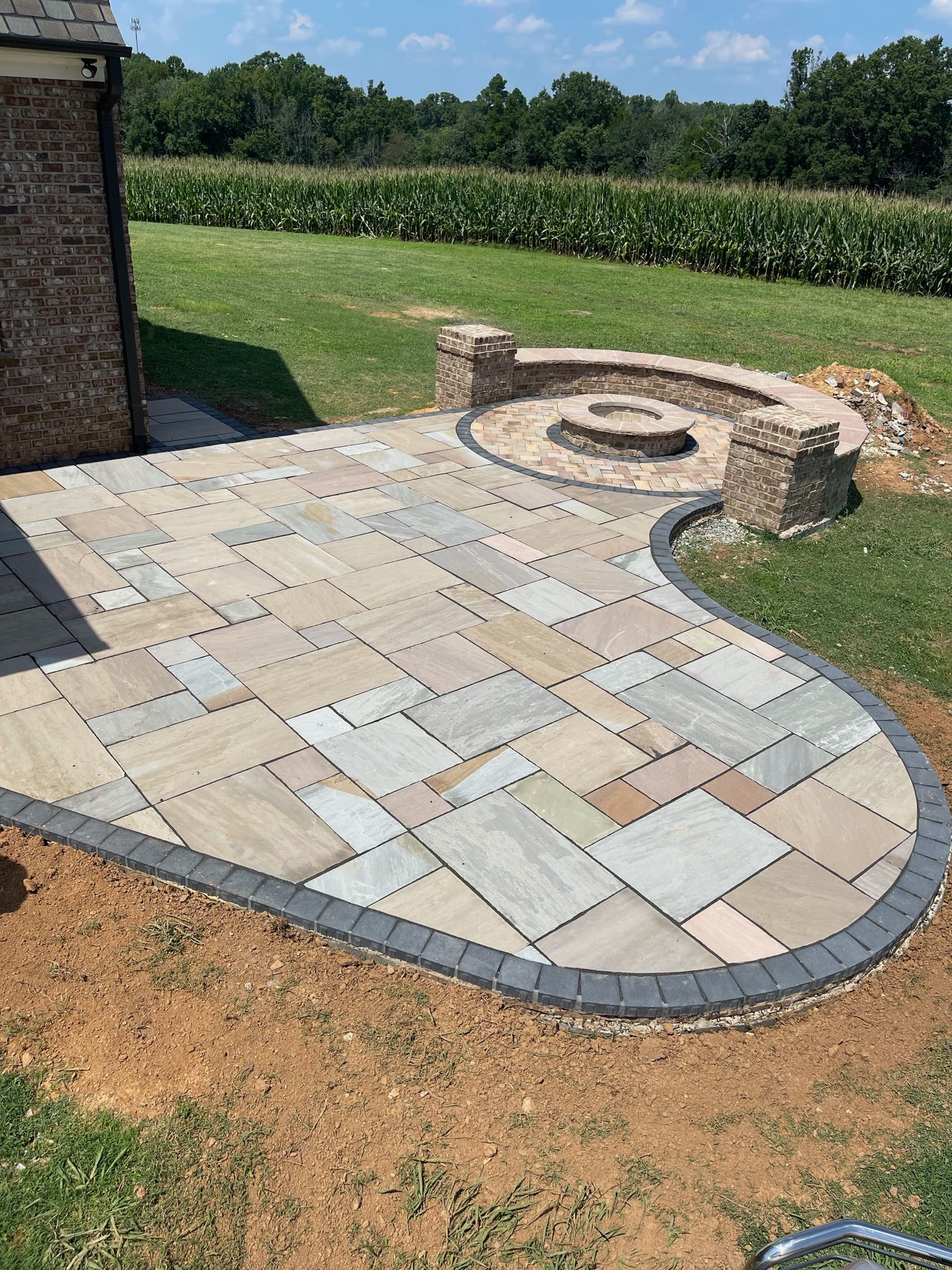 Stone patio with a fire pit, bordered by dark pavers, and a field backdrop.