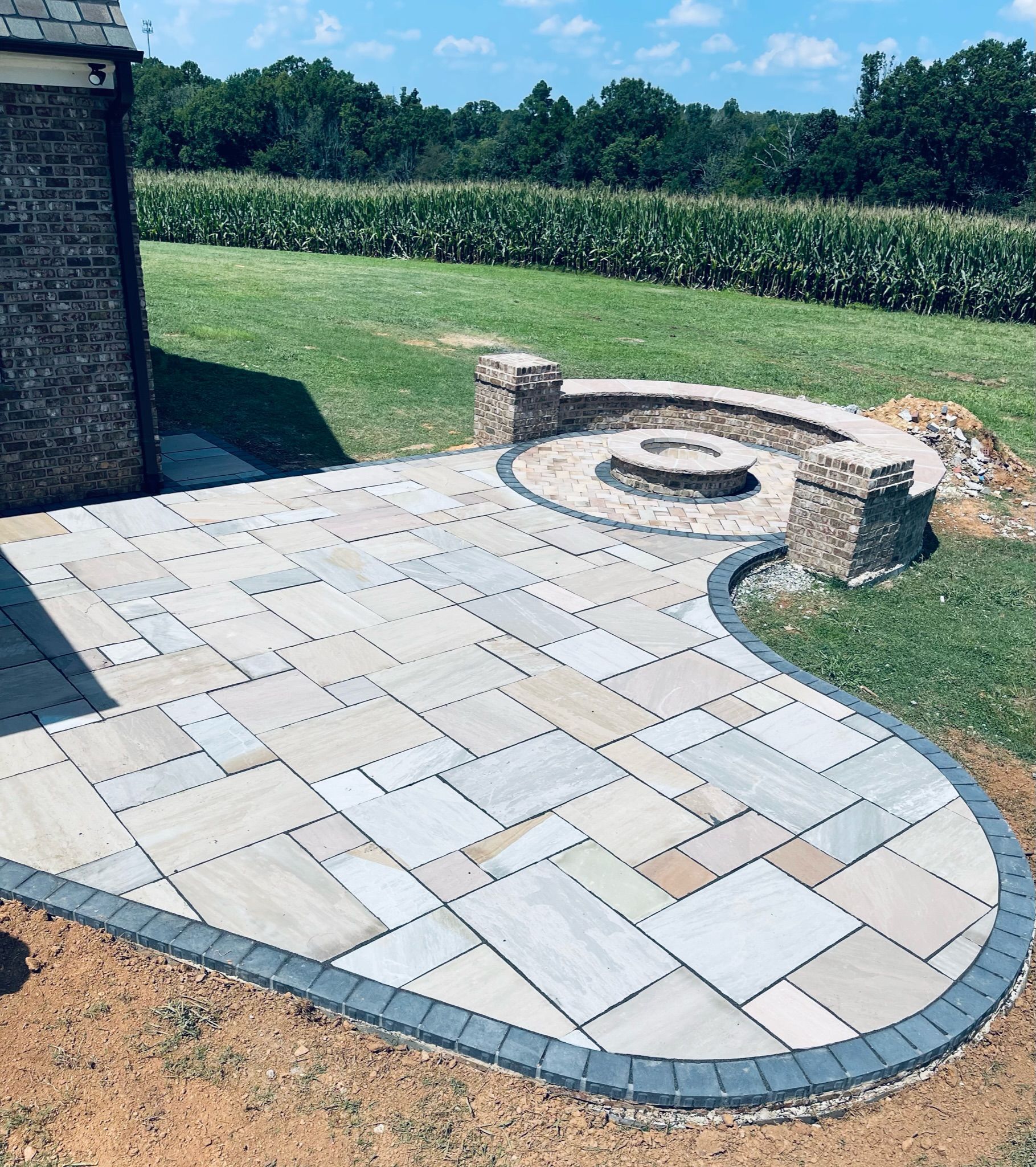 Stone patio with fire pit, grass, and brick wall against a field under a blue sky.
