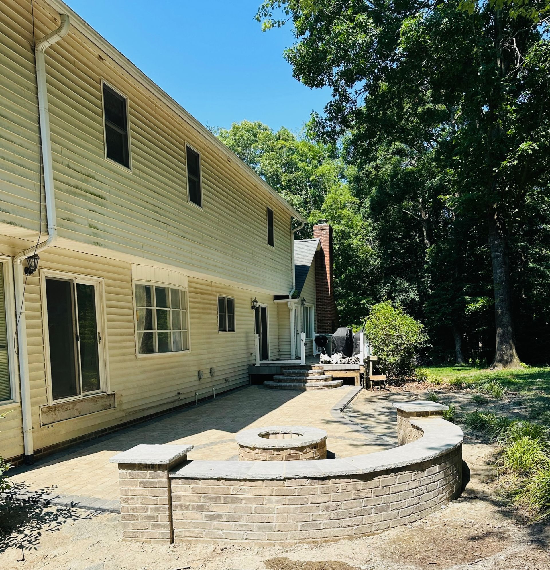 Backyard with a brick fire pit, patio, and two-story house with cream siding, under a blue sky.