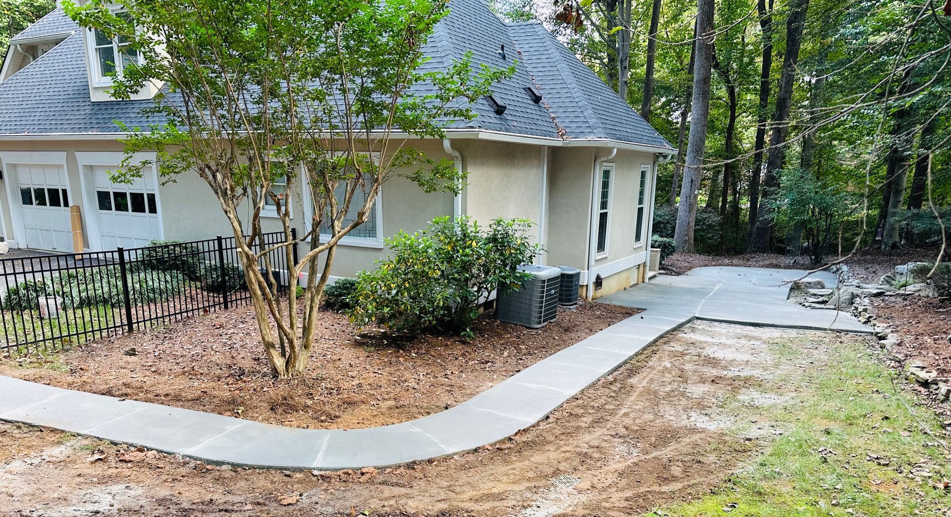 A concrete walkway leads to a light tan house with black roof, set amidst trees and mulch.