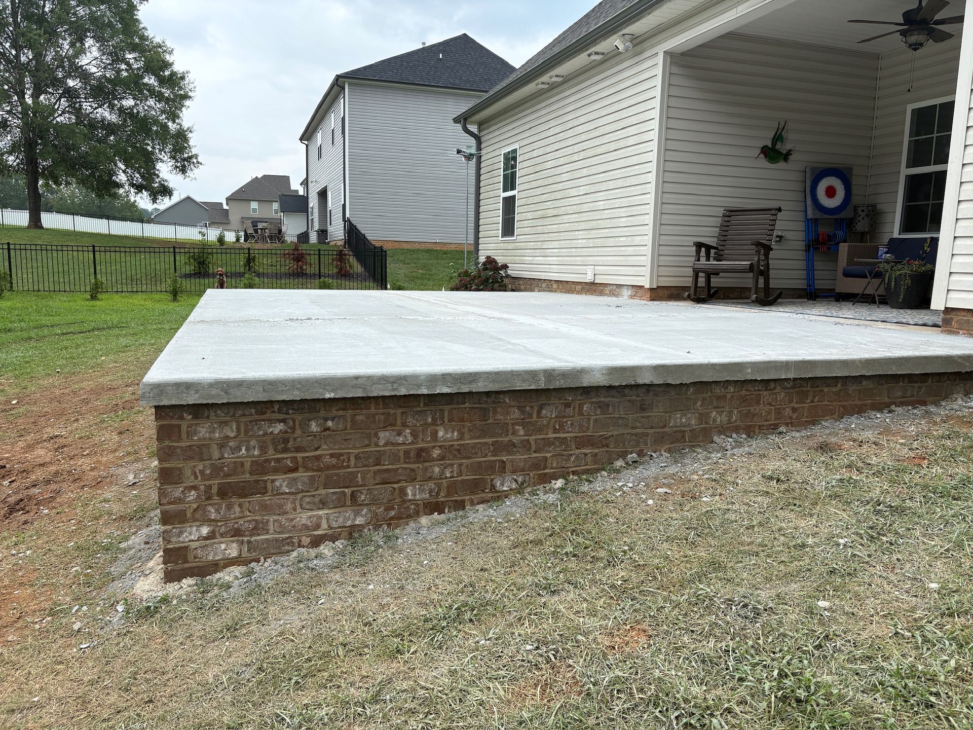 Brick-faced patio with a concrete surface. Back of a house is visible, along with a grassy yard and a fence.