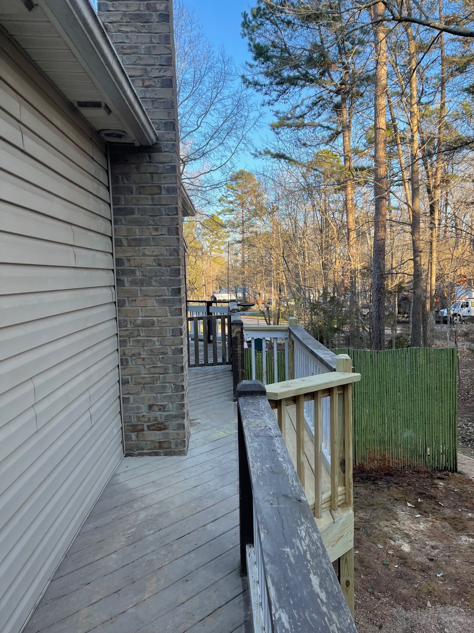 Side view of house with weathered wooden deck, brick chimney, and green fence, trees in background.