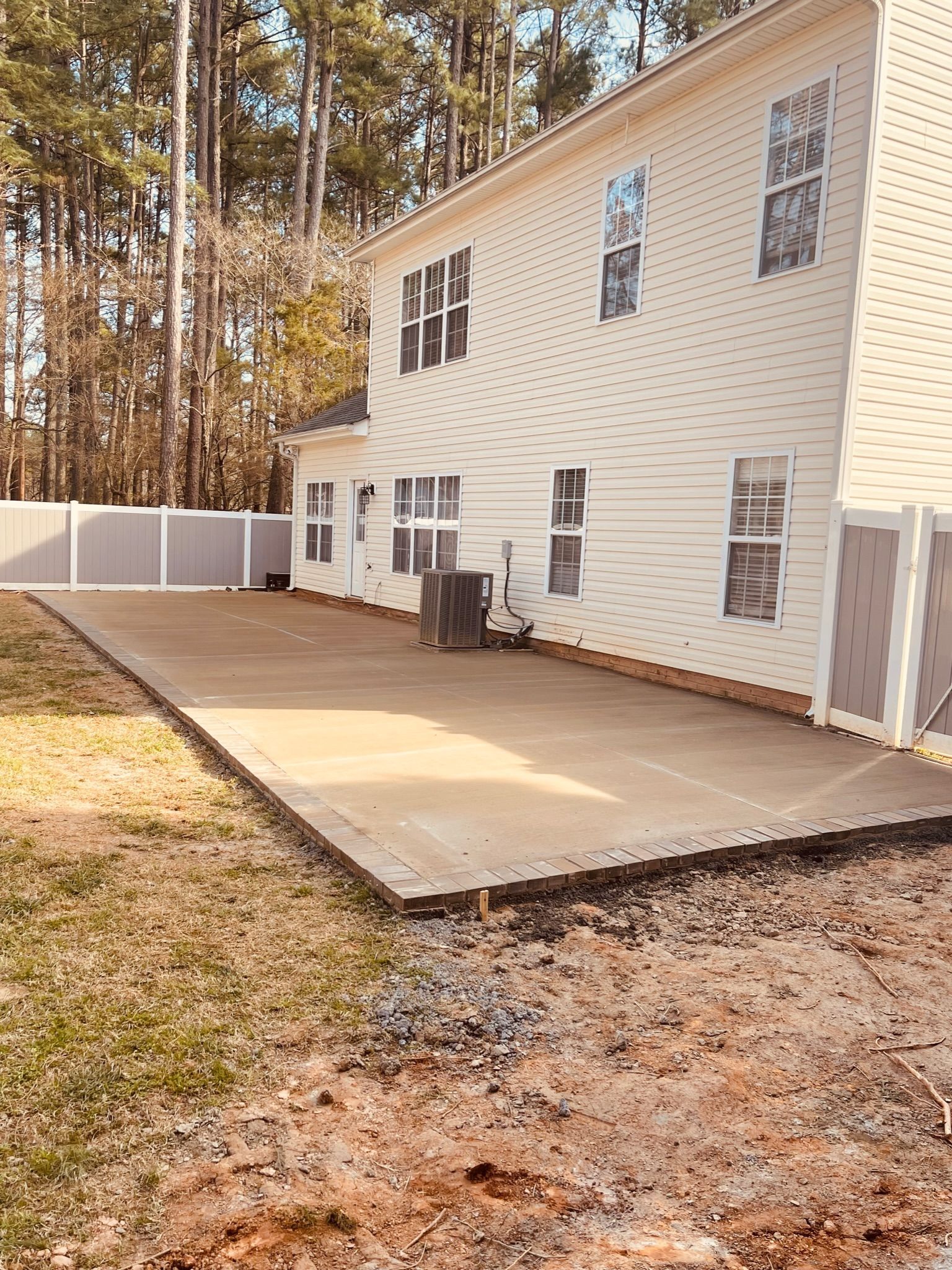 Concrete patio outside a two-story cream house with a white fence. Backyard with trees in the background.