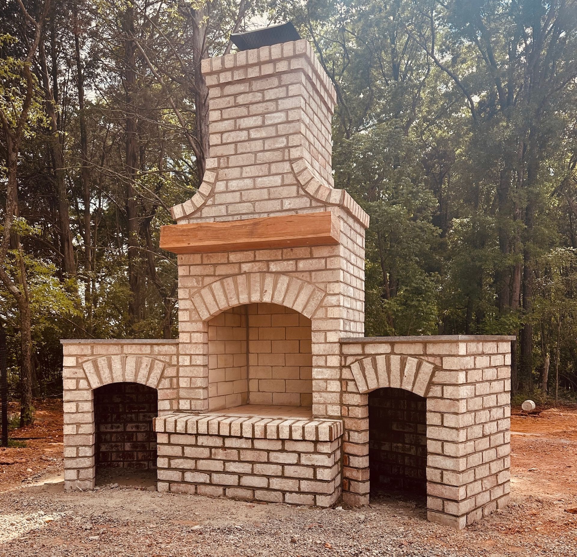 Brick outdoor fireplace with arched openings and chimney; set in a wooded area.