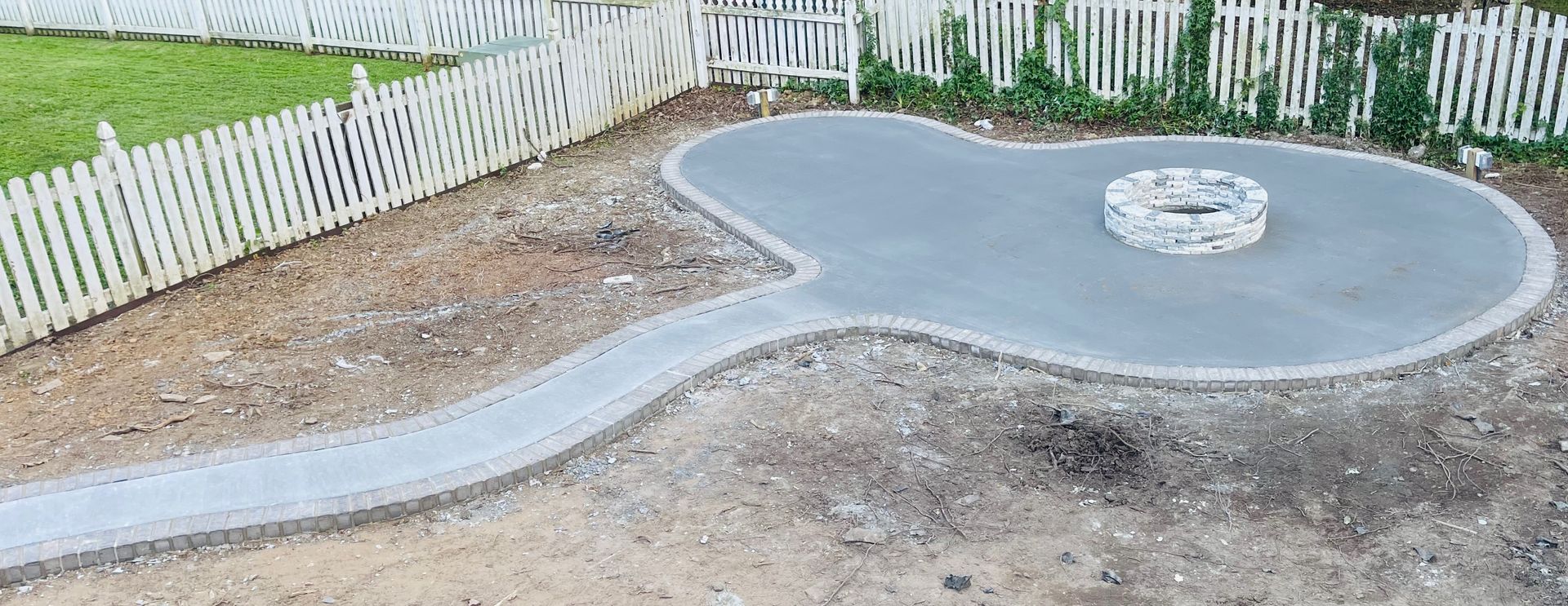 A guitar-shaped patio with a fire pit, a concrete pathway, and white picket fences.