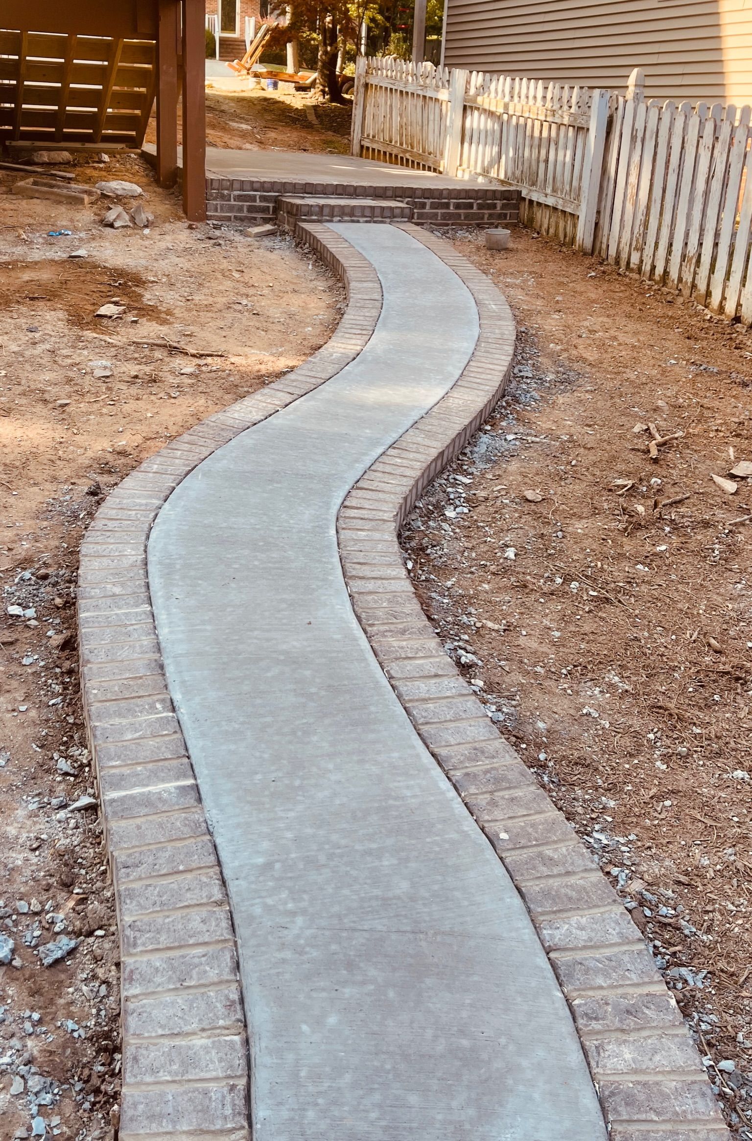 Concrete pathway with brick edging, winding through a yard, leading to steps and a wooden structure.