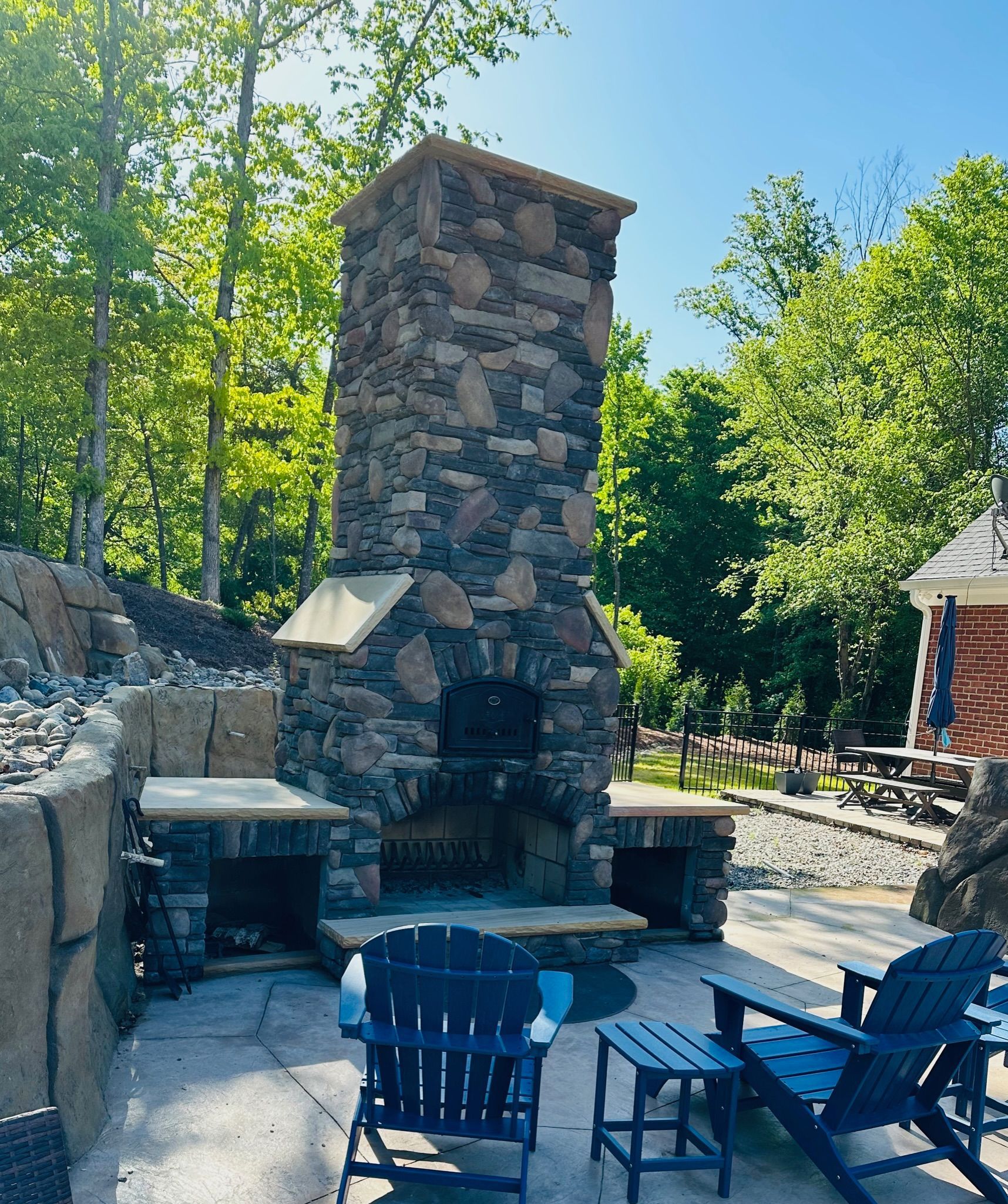 Outdoor stone fireplace with seating on a patio. Trees in the background. Blue sky.