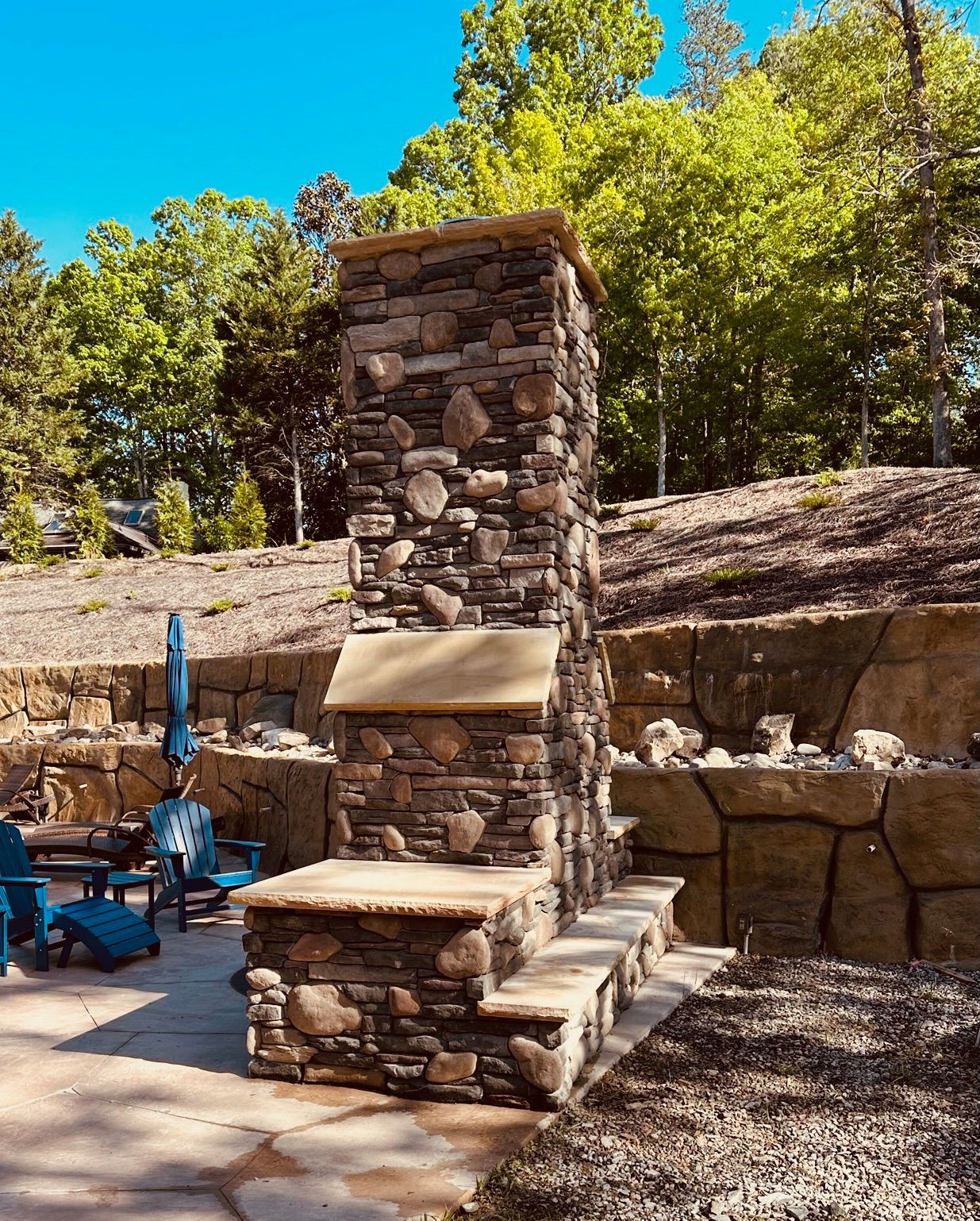 Stone outdoor fireplace on a patio, blue chairs, and trees in the background.