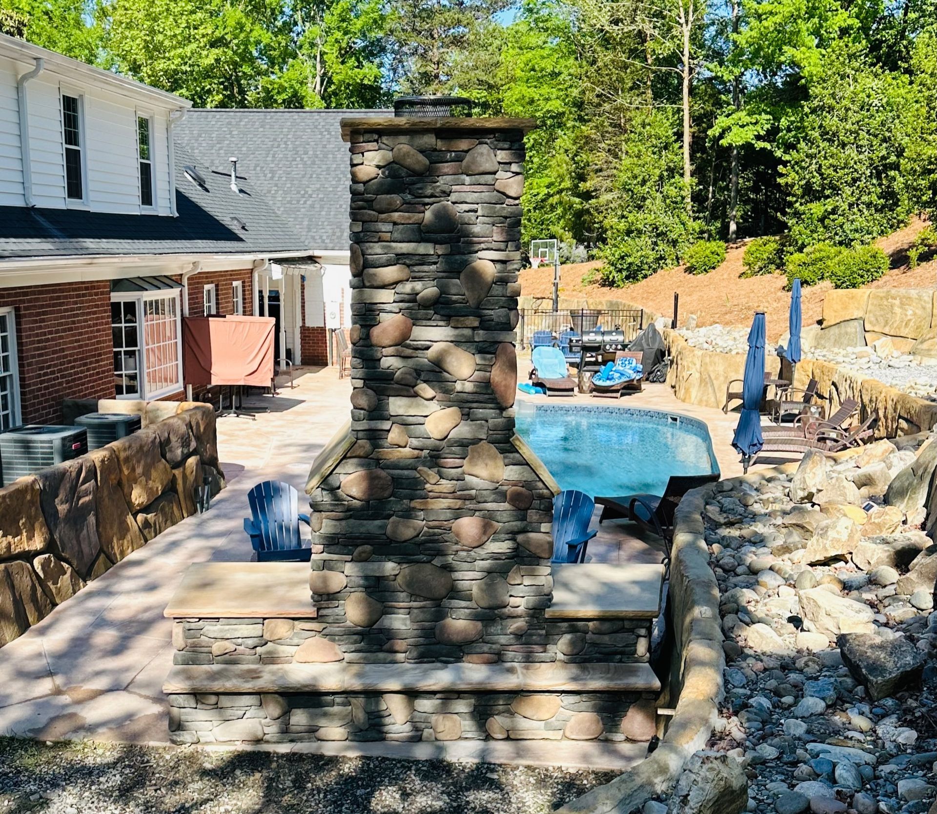 Stone fireplace on patio overlooking a pool and backyard, with trees.