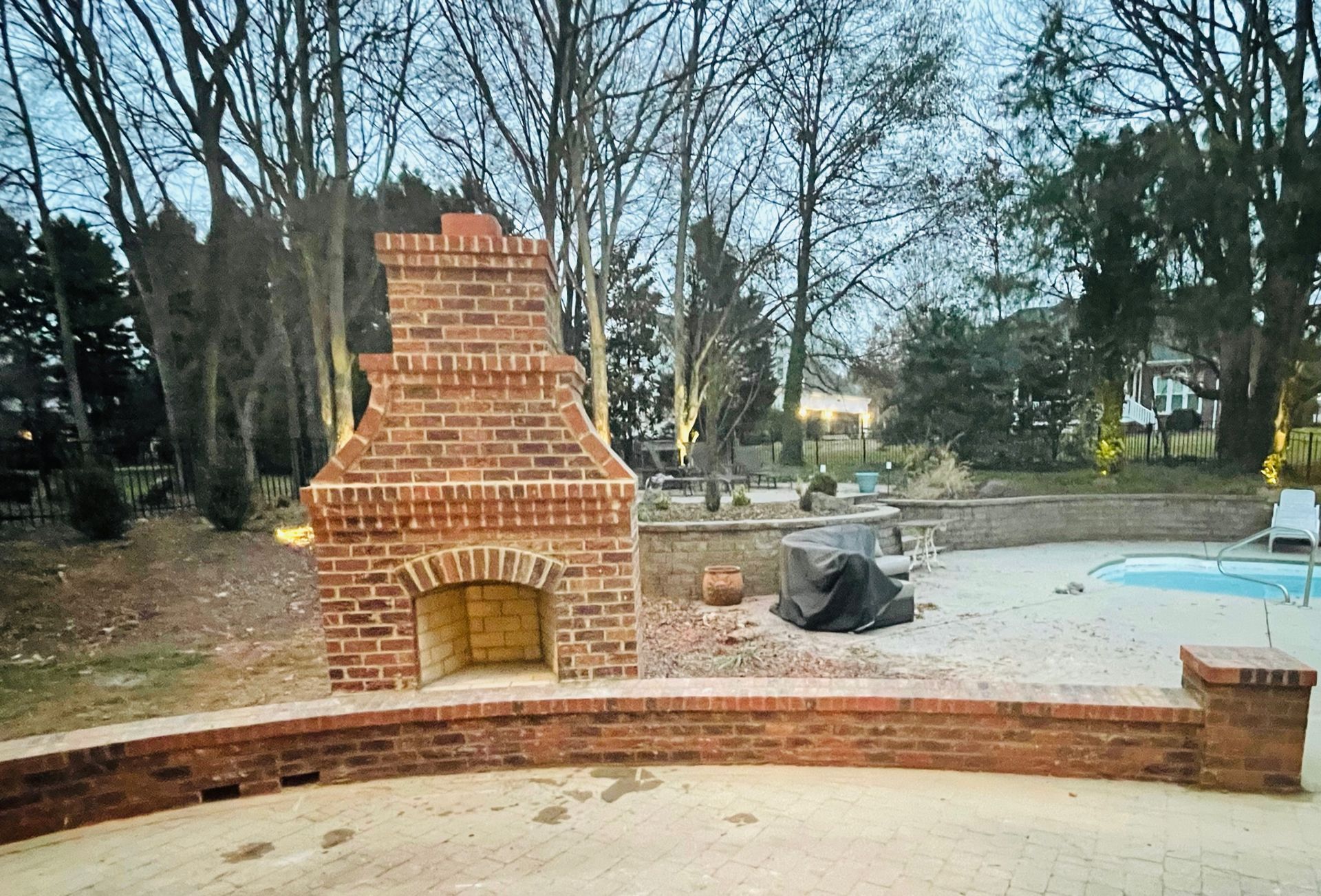 Brick outdoor fireplace with arched opening, brick wall, pool, and trees in background.