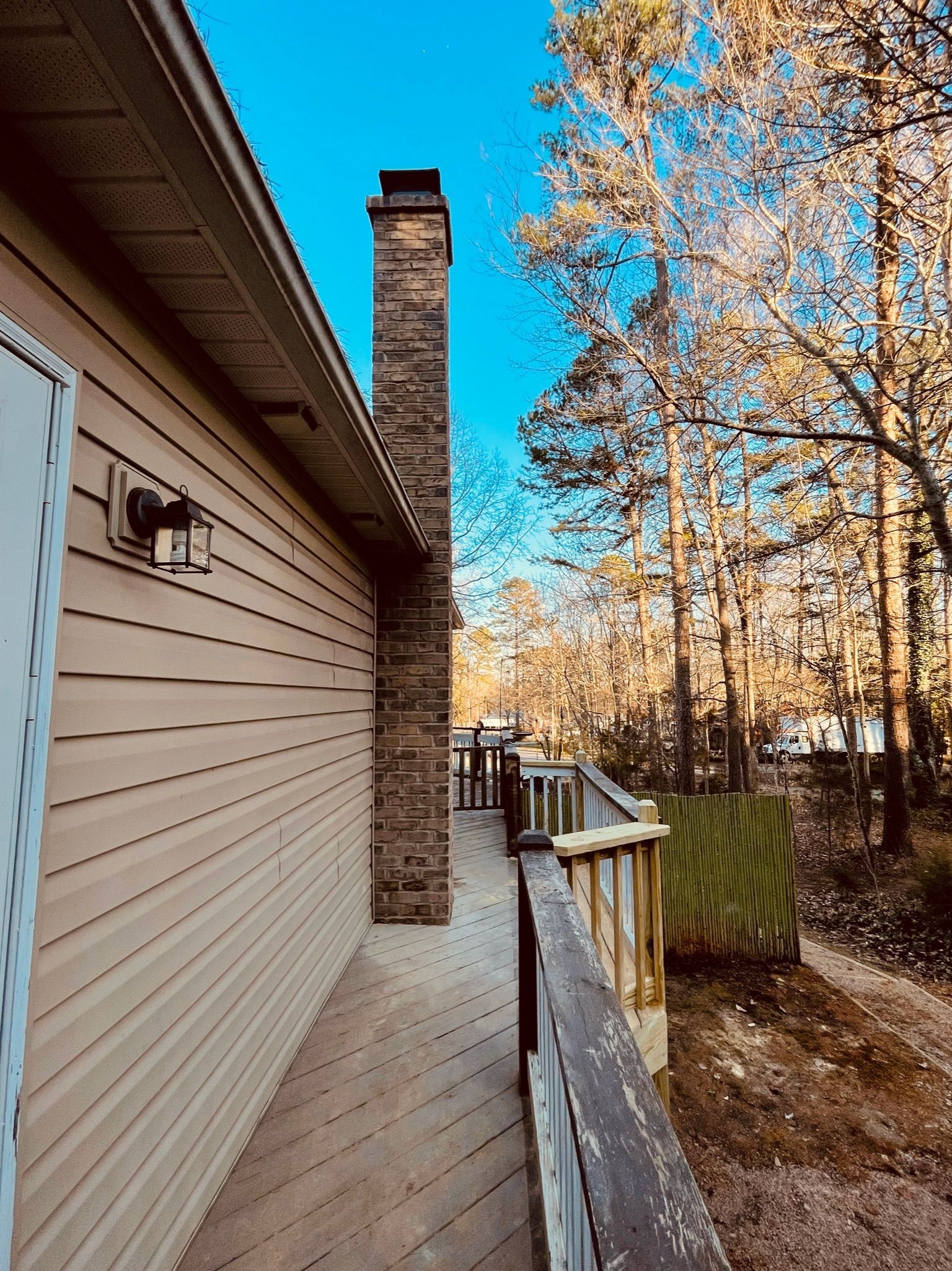 Side of a house with wooden deck, brick chimney, and trees in background, under a blue sky.