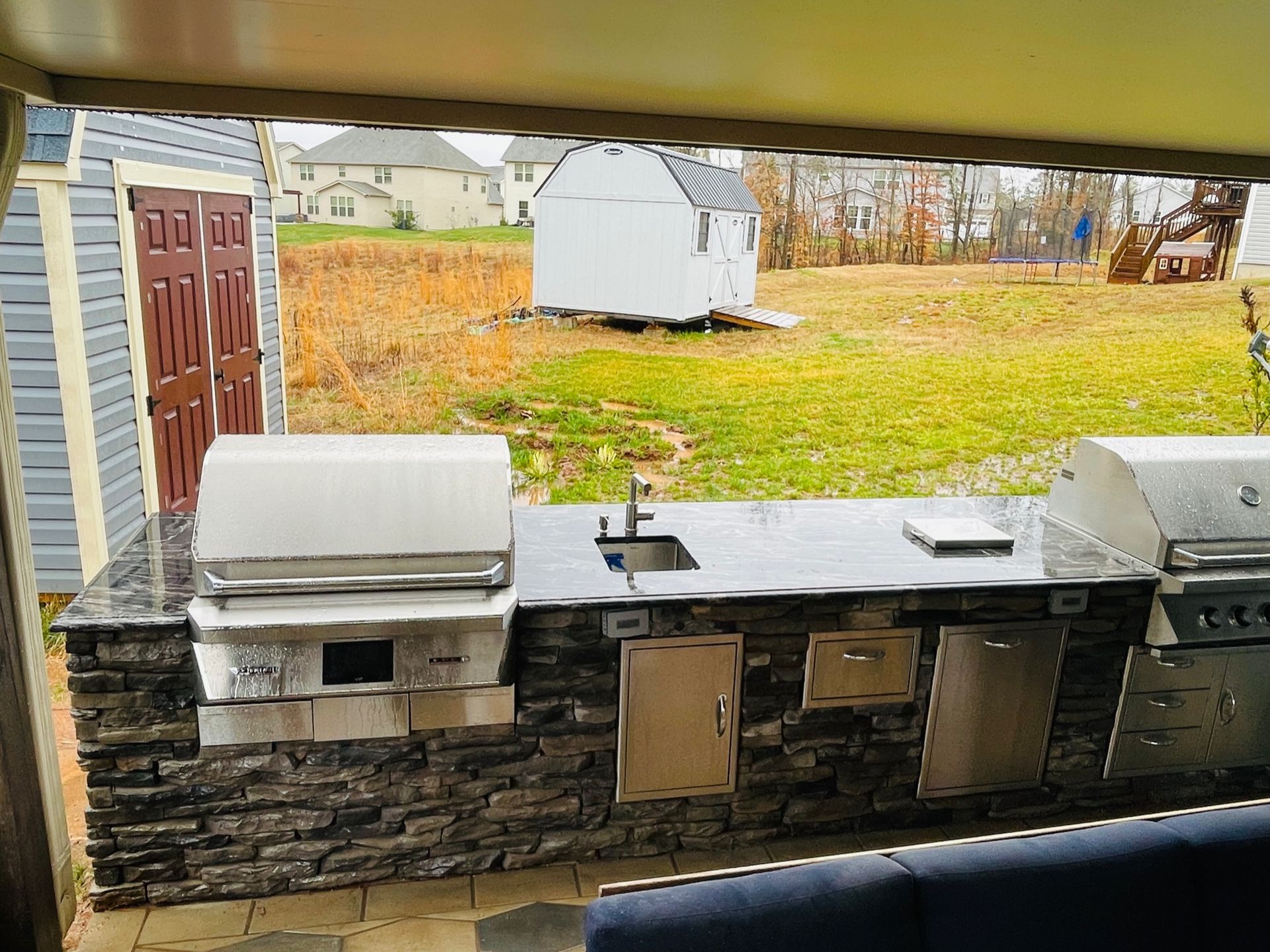 Outdoor kitchen with stone facade, stainless steel appliances, sink, in a backyard setting.