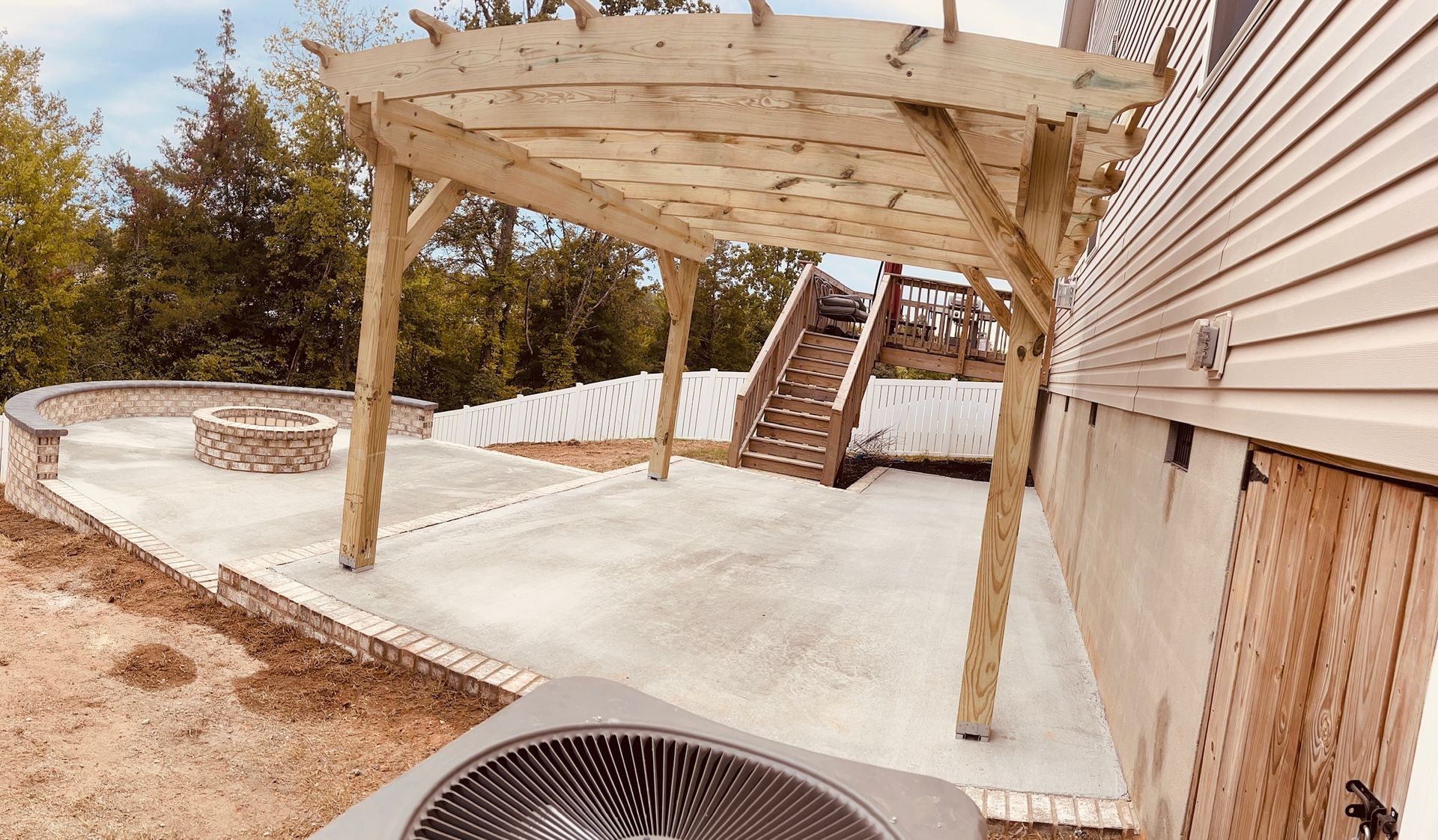 Wooden pergola over a concrete patio, next to a house with stairs.