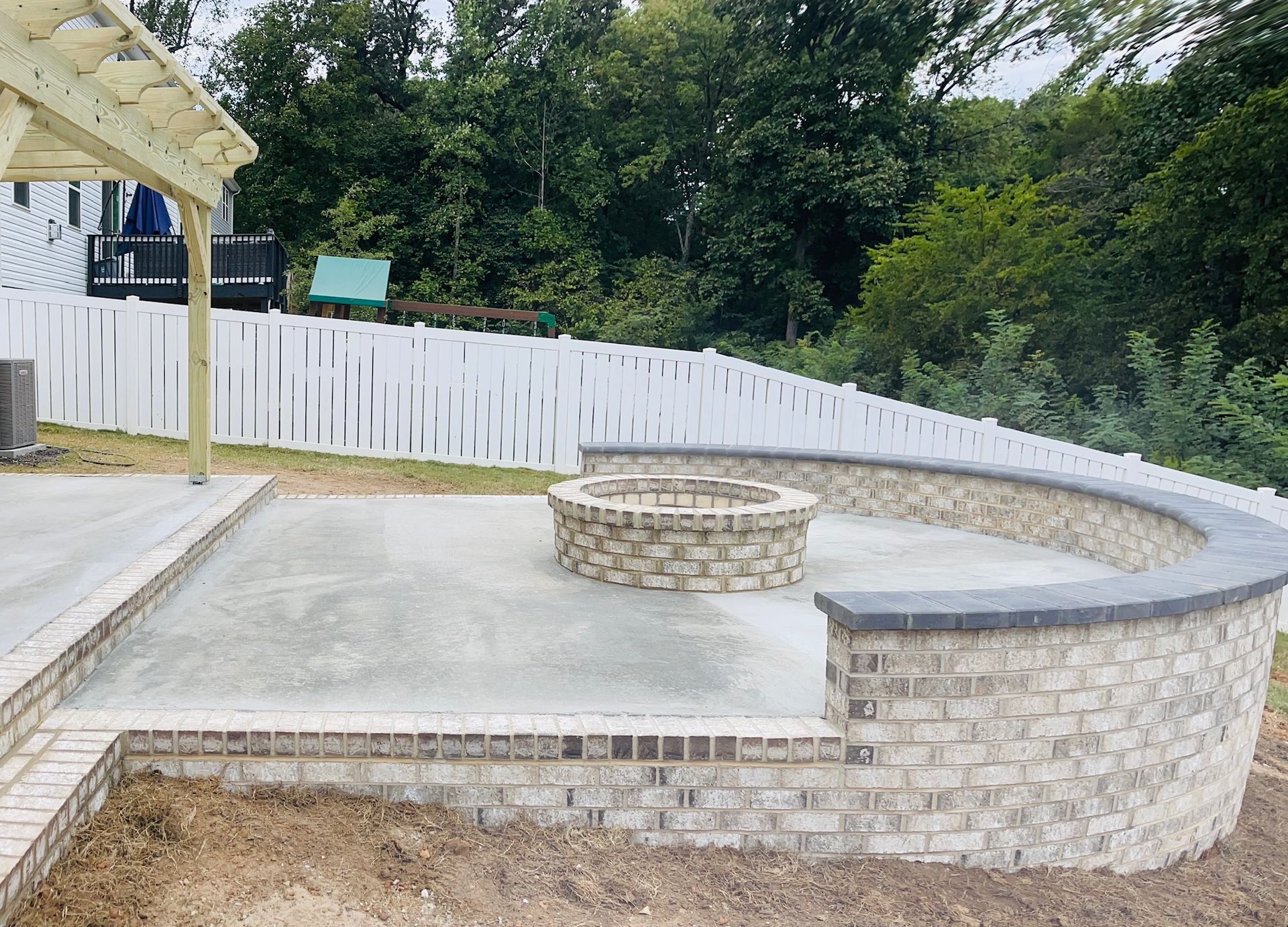A brick patio with a fire pit and curved seating, set against a white picket fence and trees.