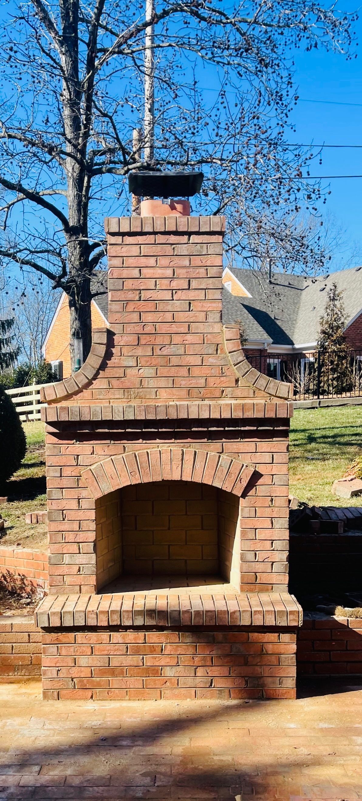 Brick outdoor fireplace with arched opening, chimney, and black cap, set against a blue sky and trees.