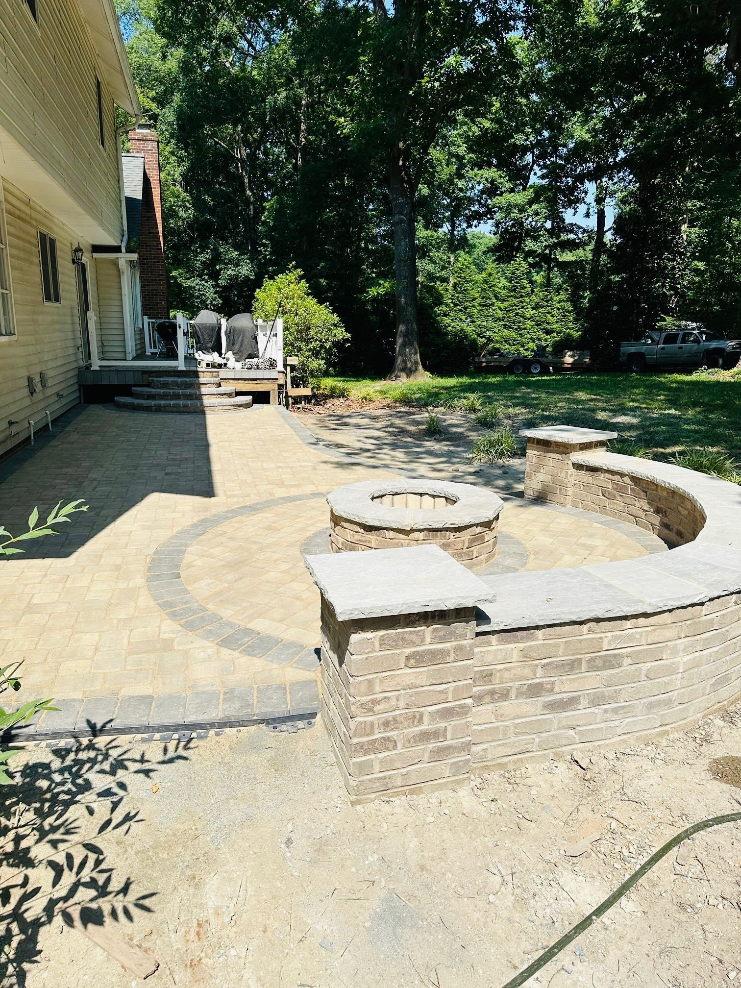 Backyard patio with fire pit, brick wall, and landscaping. Beige pavers, lush greenery.