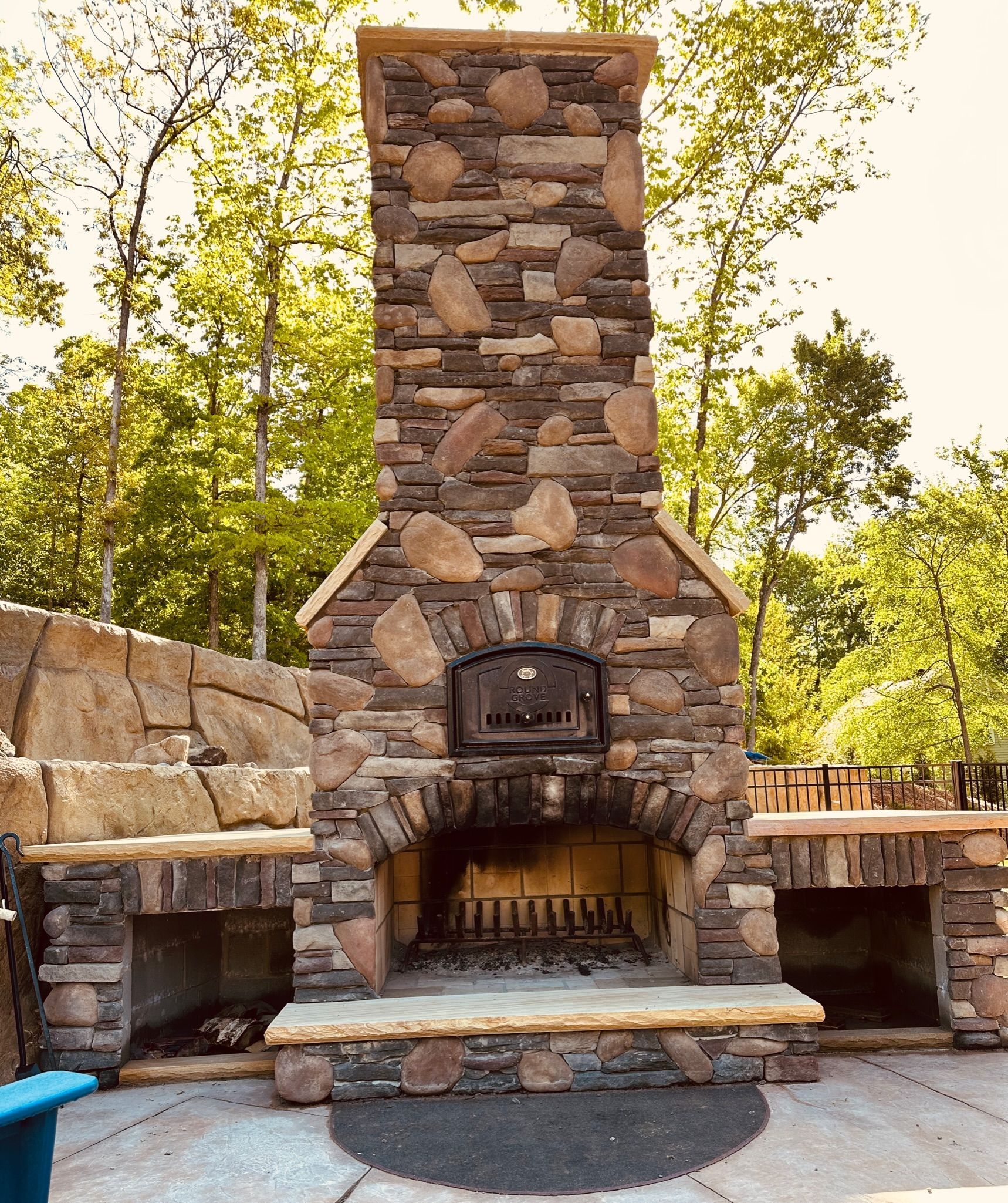 Outdoor stone fireplace with a built-in oven, surrounded by stone counters on a patio, trees in the background.